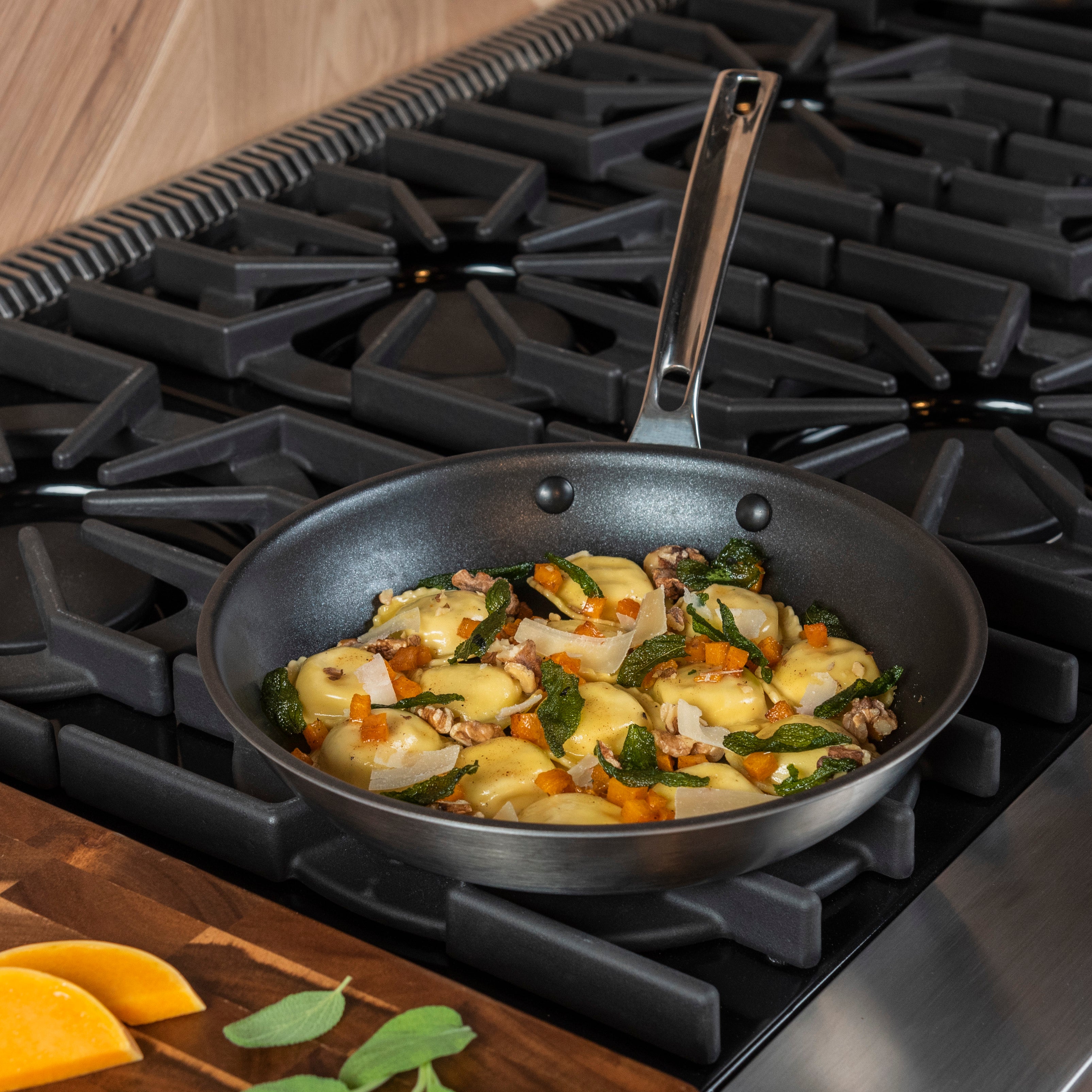 A skillet with sautéed vegetables, herbs, and possibly melted cheese sits on a gas stove. Nearby, a wooden cutting board holds orange slices and fresh sage leaves.