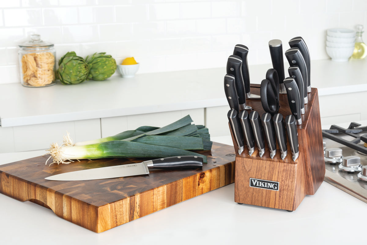A wooden knife block set with various knives and scissors sits on a kitchen counter. Next to it, a leek and a chef’s knife rest on a wooden cutting board. Artichokes and pasta in jars are in the background.