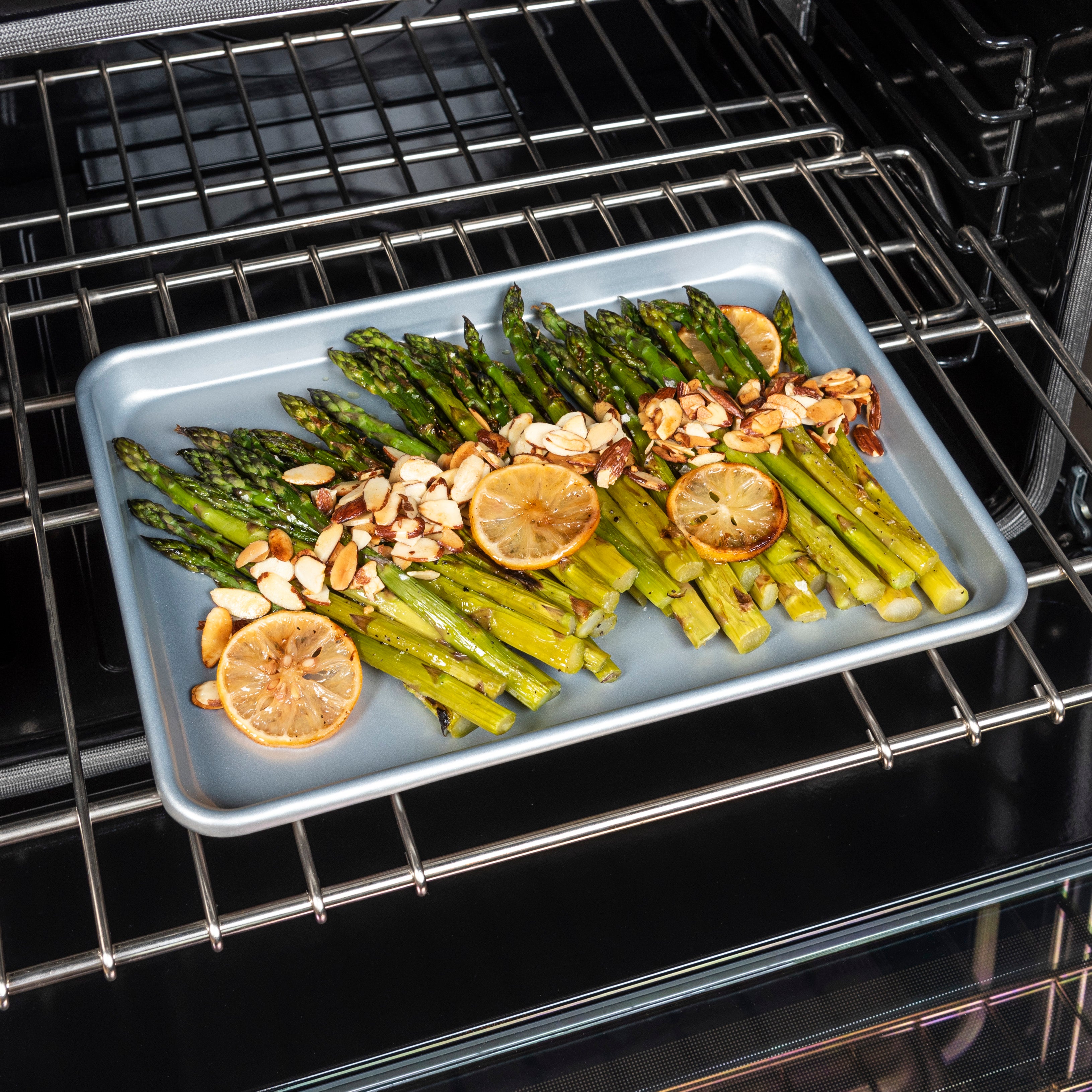 A tray of roasted asparagus topped with sliced almonds and lemon slices sits on an oven rack, ready to be cooked.