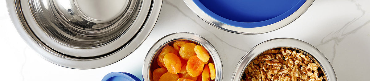 Four stainless steel containers, two with blue lids, hold dried apricots and chopped nuts. The containers are arranged on a white marble surface.