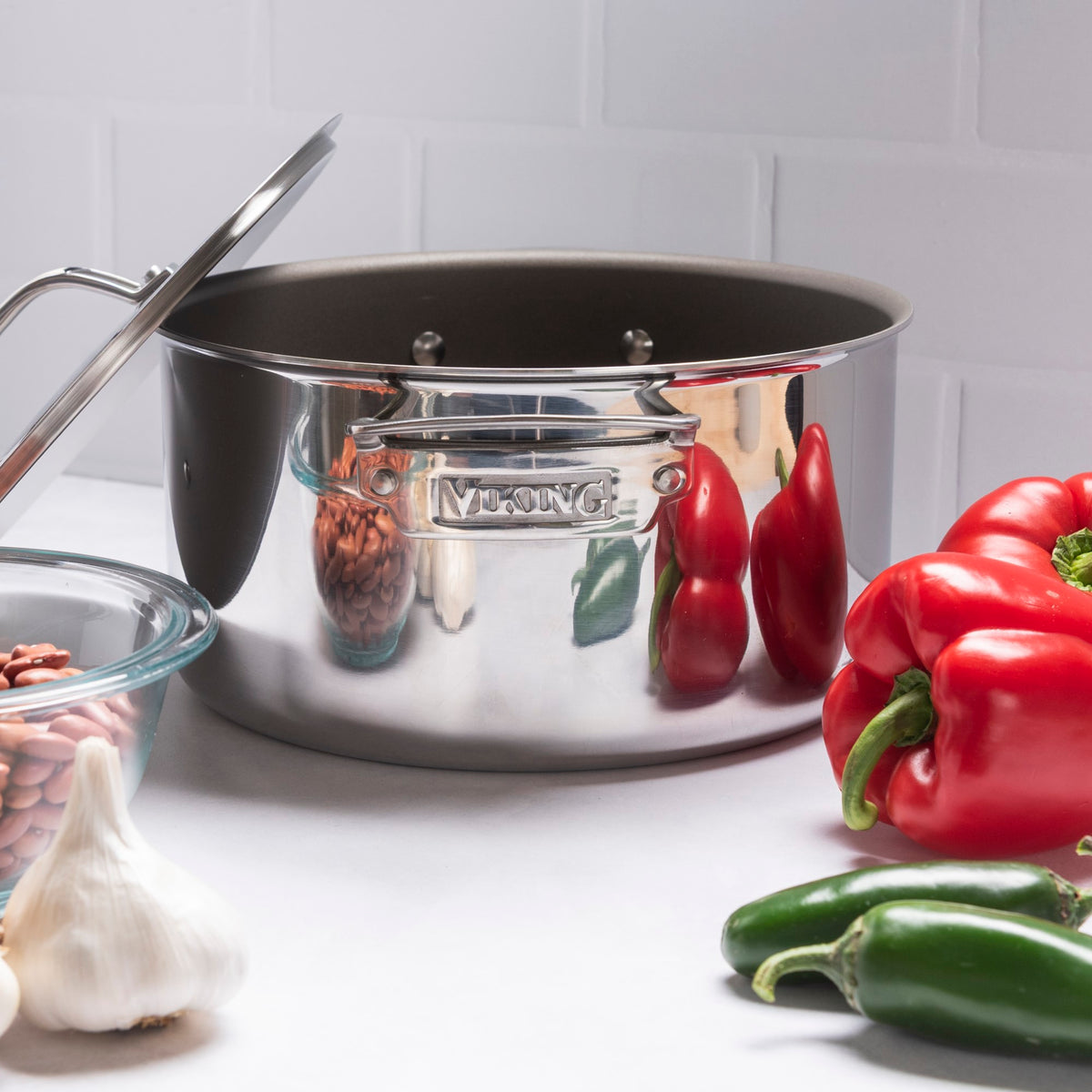 The Viking Culinary Products PureGlide Pro 3-Ply 6-Quart Dutch Oven with Metal Lid sits on a counter amid red bell peppers, jalapeños, garlic, and beans, set against a white tiled background.