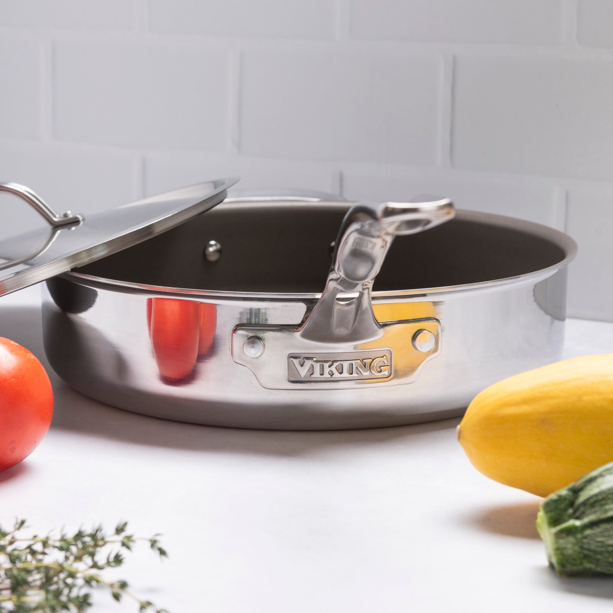 A Viking PureGlide Pro 3-Ply 4.5-Quart Saute Pan with Metal Lid by Viking Culinary Products sits on a white countertop with tomato, yellow squash, zucchini, and thyme—ideal for healthy cooking—against a white tiled backdrop.