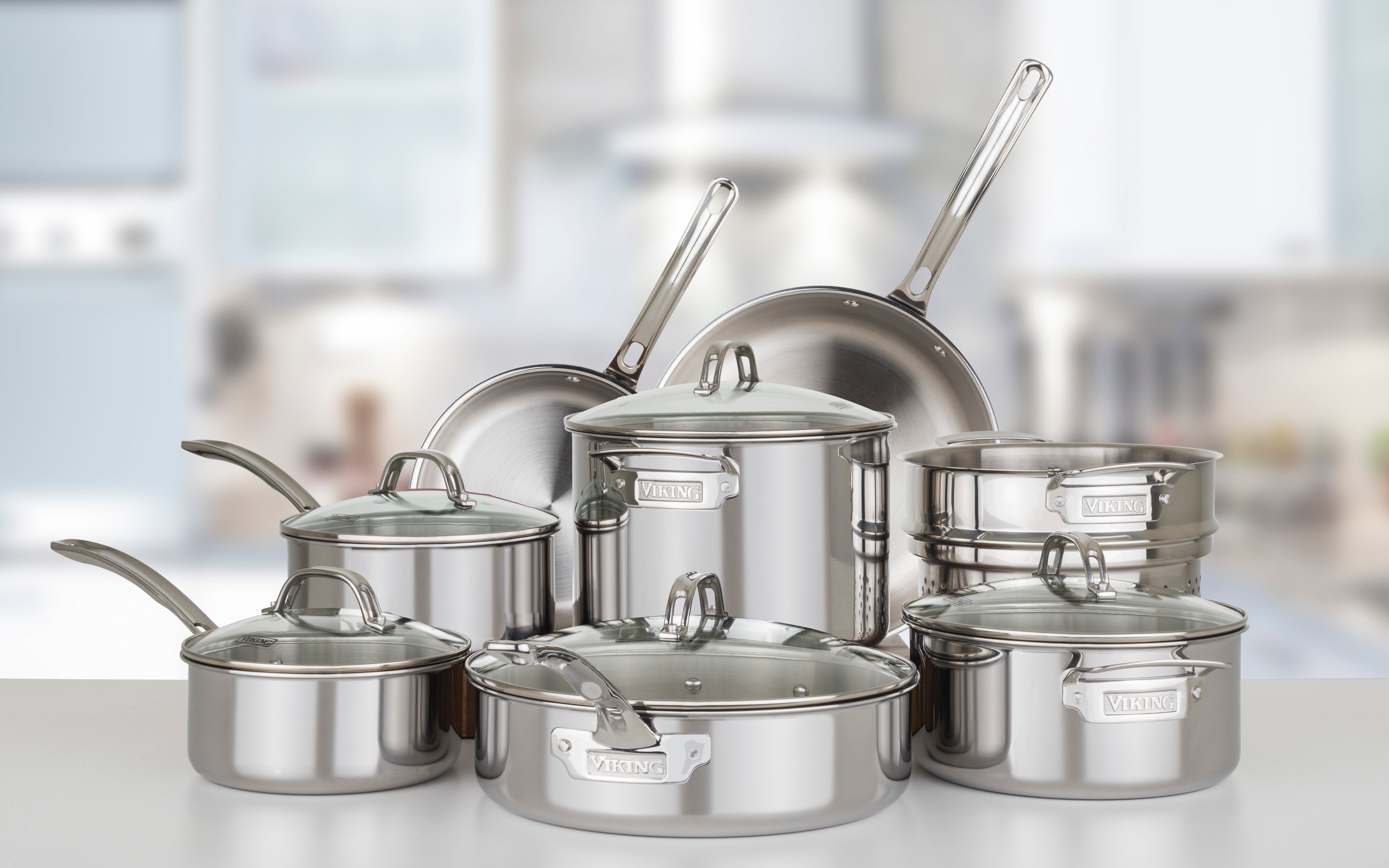 The Viking Culinary Products 3-Ply Stainless Steel 13-Piece Cookware Set with glass lids is neatly arranged on a kitchen counter, with a blurred kitchen background. All pieces are dishwasher safe.