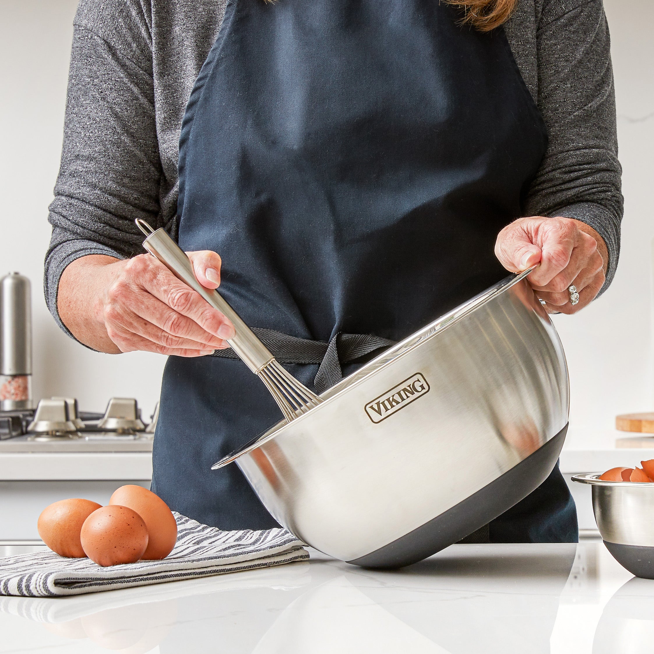A person wearing a dark apron whisks ingredients in a Viking Culinary Products 9-Piece Stainless Steel Mixing Bowl with Strainer and black silicone base, on a kitchen counter with eggs and a striped towel nearby.