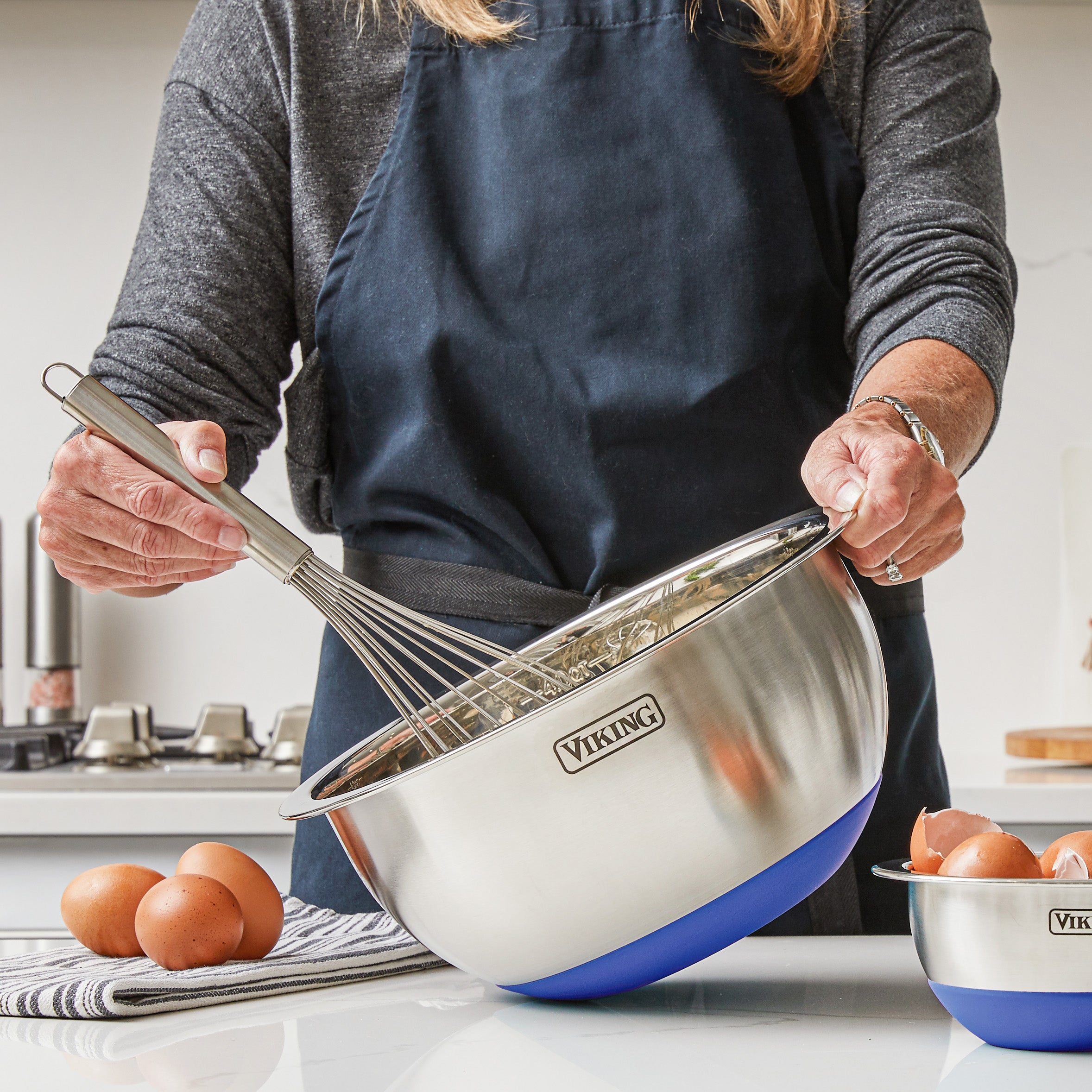 A person in a dark apron whisks ingredients in a Viking Culinary 9-Piece Stainless Steel Mixing Bowl with Blue Silicone Base. Brown eggs rest nearby on a striped cloth and in a smaller bowl on the kitchen counter.