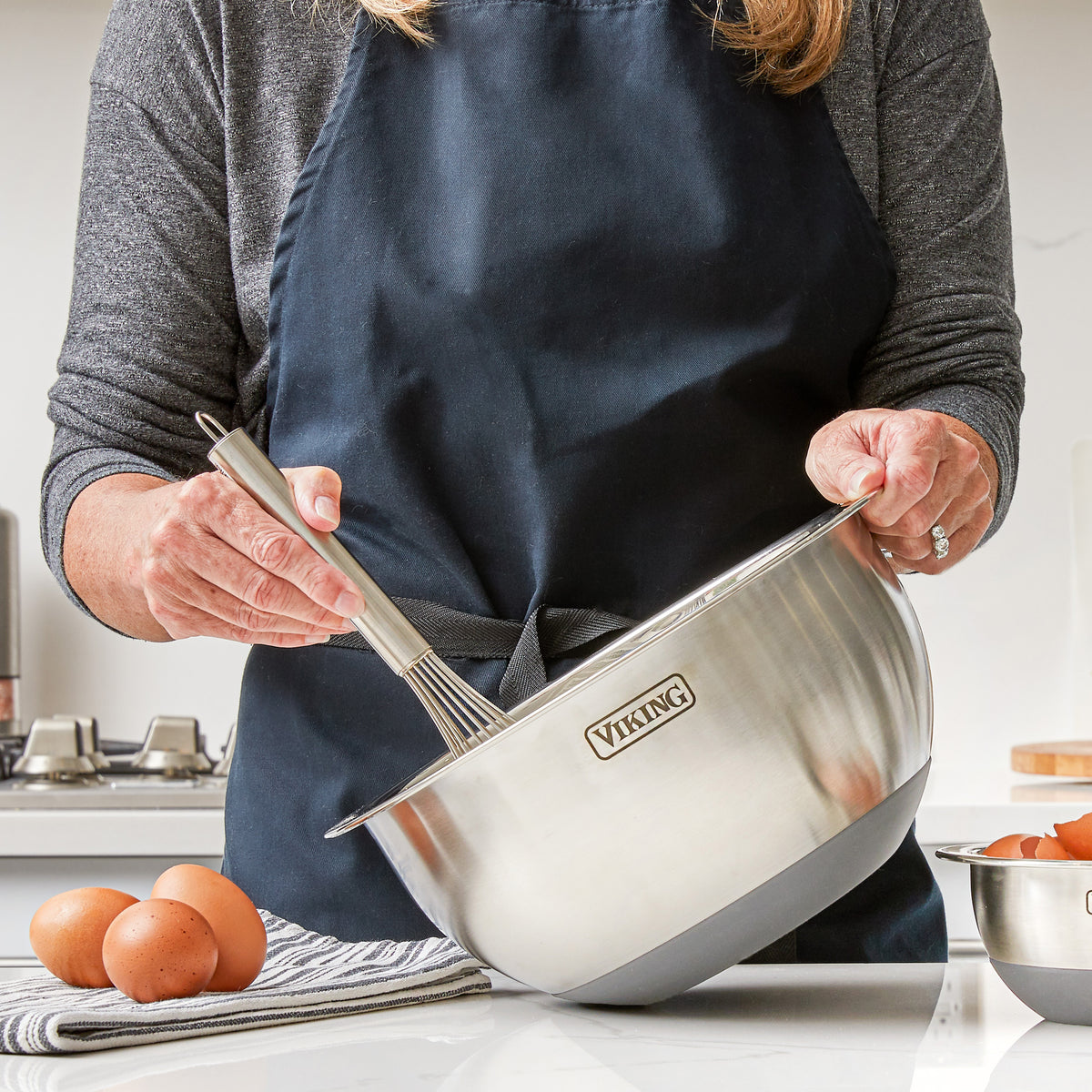 Wearing a dark apron, a person whisks ingredients in a Viking 9-Piece Stainless Steel Mixing Bowl Set with Strainer by Viking Culinary Products—ideal for anyone who values mixing bowls designed for compact storage.