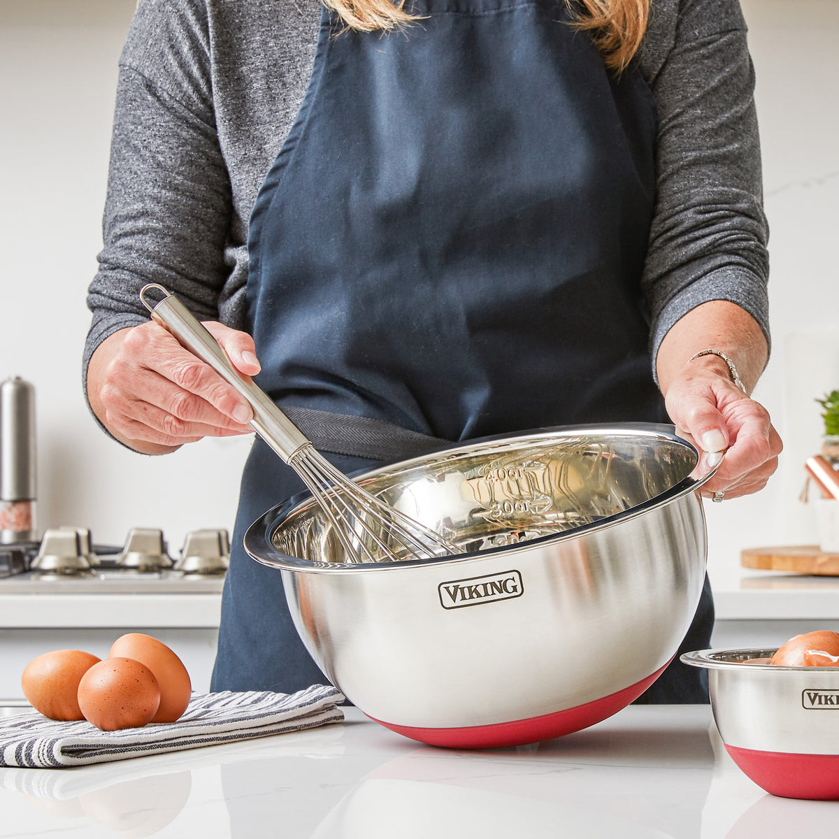 A person in a navy apron holds a Viking 9-Piece Stainless Steel Mixing Bowl Set with Strainer, Red by Viking Culinary Products—ideal for those seeking mixing bowls designed for compact storage. Eggs, a towel, and another bowl rest on the kitchen counter.