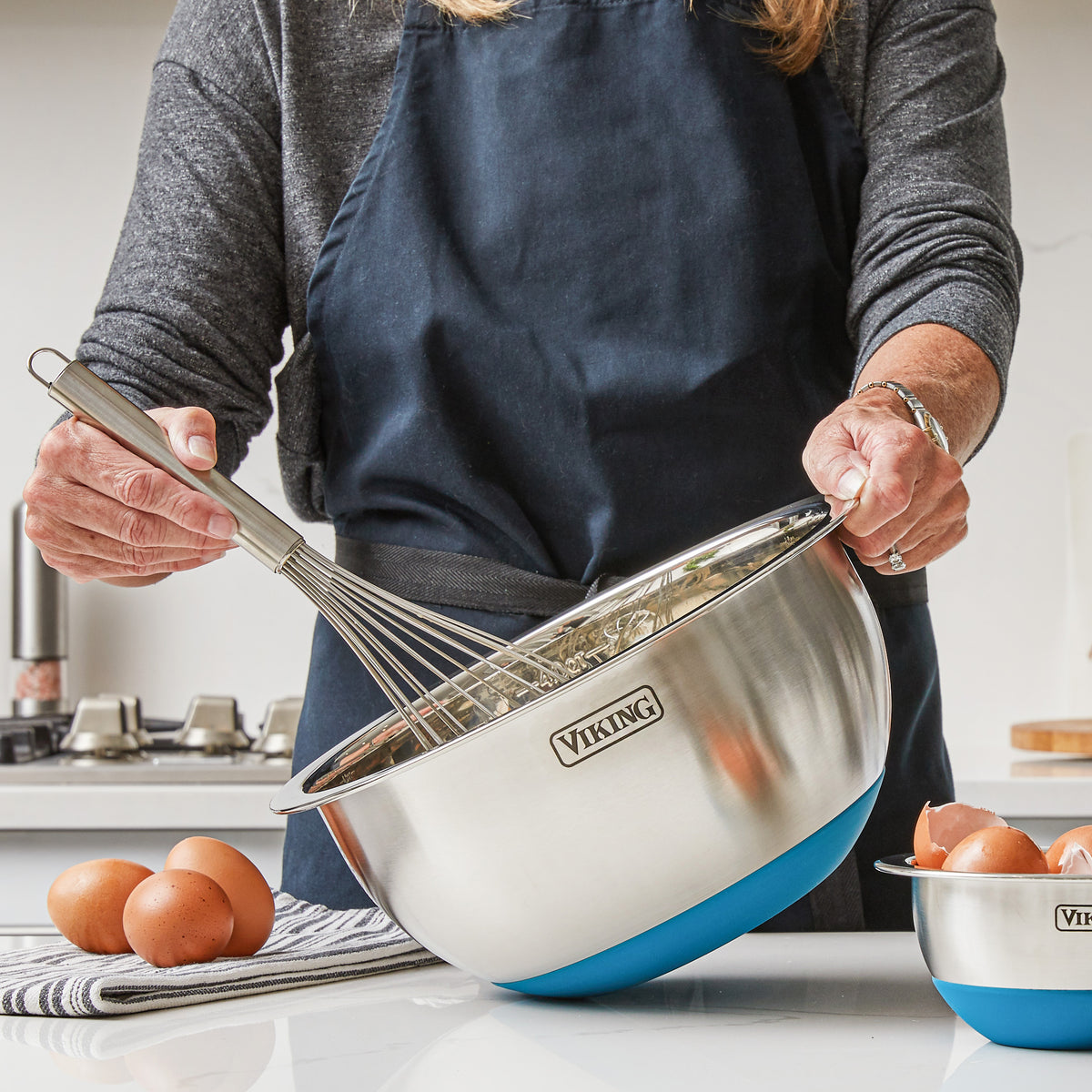 A person in a dark apron whisks ingredients in a large Viking Culinary Products 9-Piece Stainless Steel Mixing Bowl with Strainer, Teal, perfect for those who value compact storage in busy kitchens. Eggs and an empty bowl sit nearby.