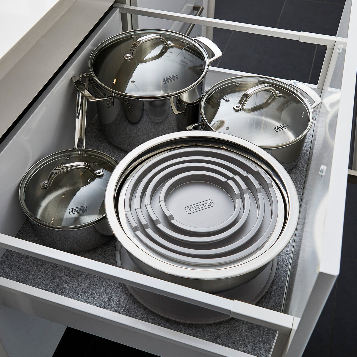 Open kitchen drawer containing three stainless steel pots with lids and a set of nested, circular, gray Viking brand mixing bowls with lids. The drawer has a gray liner, and the kitchen flooring is dark.