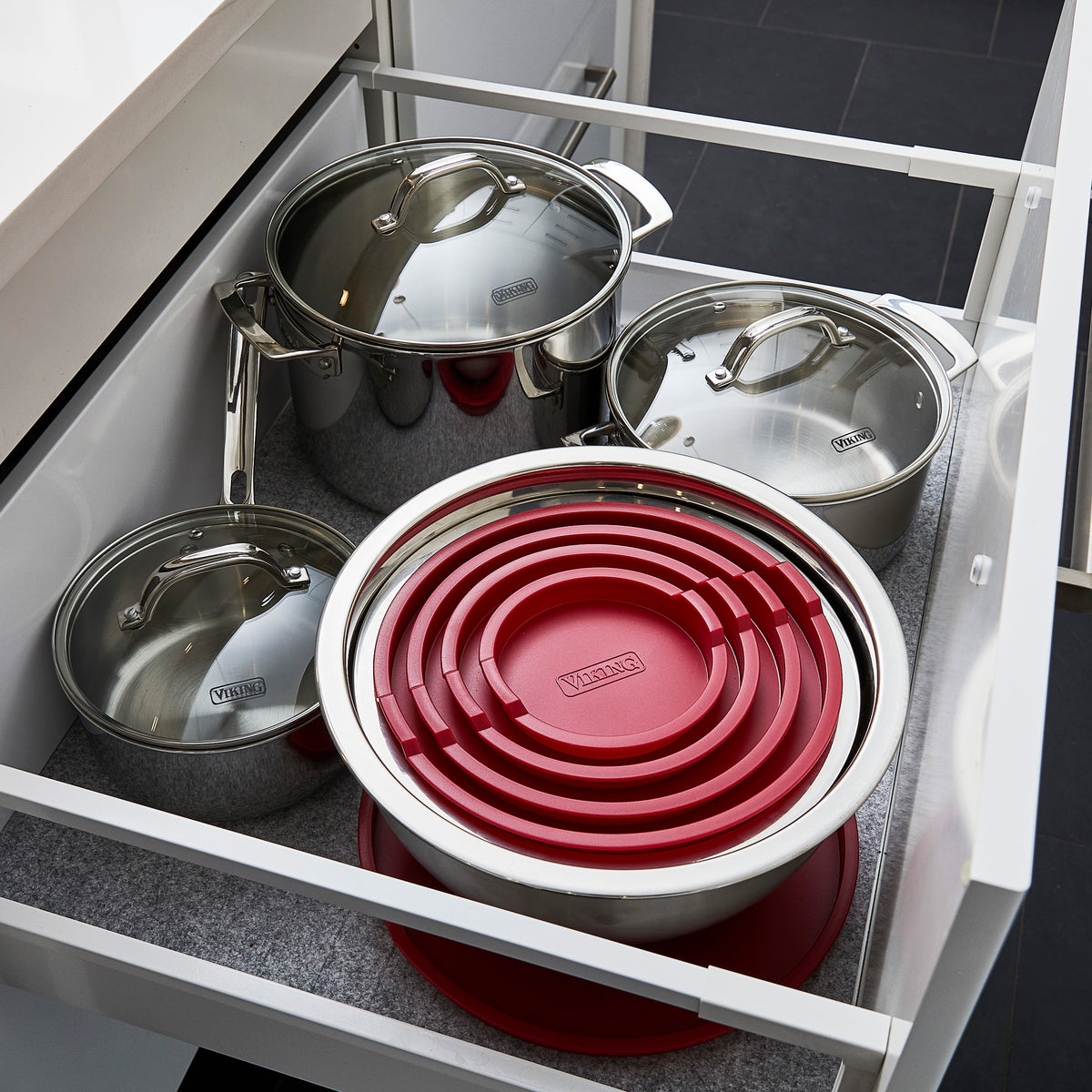 An open kitchen drawer containing stainless steel pots with lids and a set of stainless steel mixing bowls with red lids, neatly nested together. The drawer is lined with a grey mat.