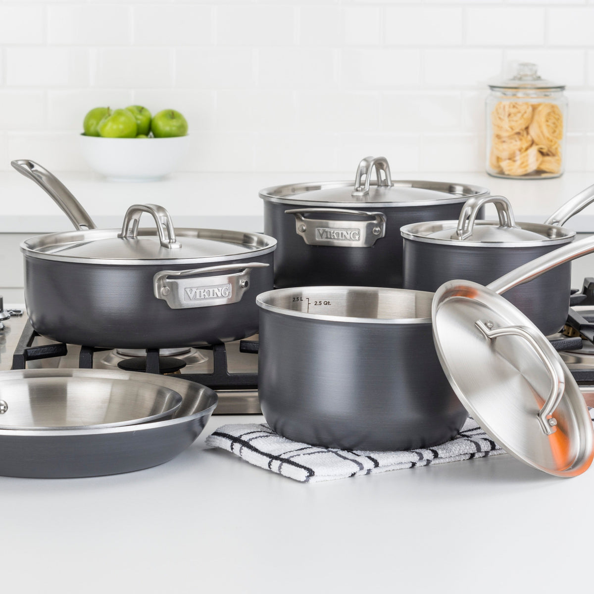 A set of stainless steel and black cookware, including pots with lids and a frying pan, is arranged on a stove and a striped towel in a modern kitchen with white subway tile and a jar of pasta in the background.