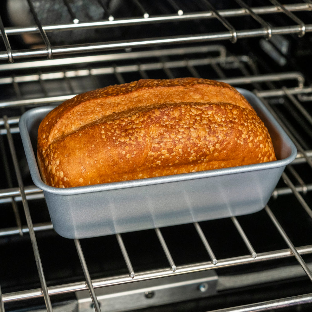 A golden-brown loaf of bread rests in a Viking Aluminized Steel Nonstick 9-inch Loaf Pan from Viking Culinary Products on an oven rack.