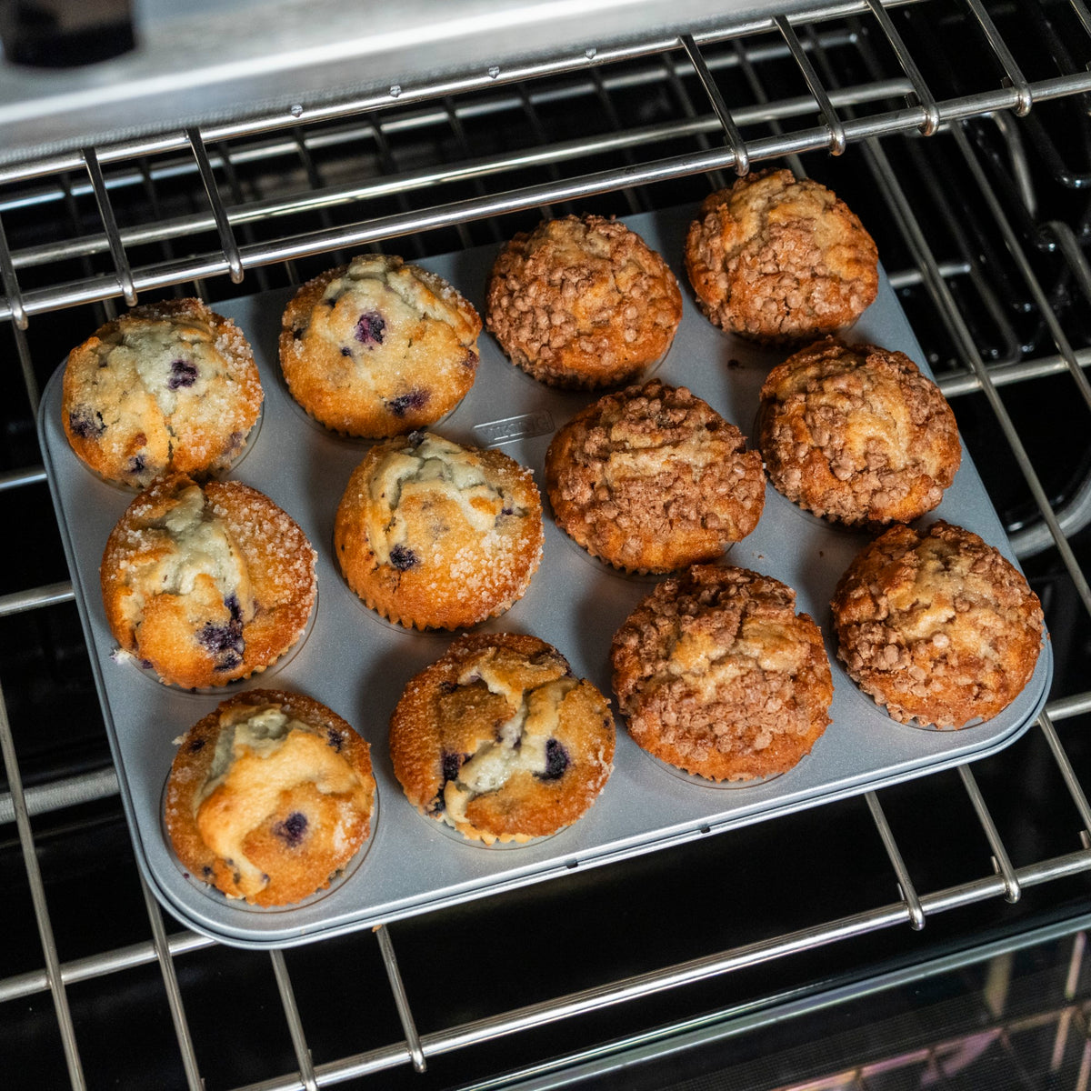 The Viking Aluminized Steel Nonstick 12-Cup Muffin Pan by Viking Culinary Products holds six blueberry and six crumb-topped muffins, all freshly baked and golden brown on the oven rack.