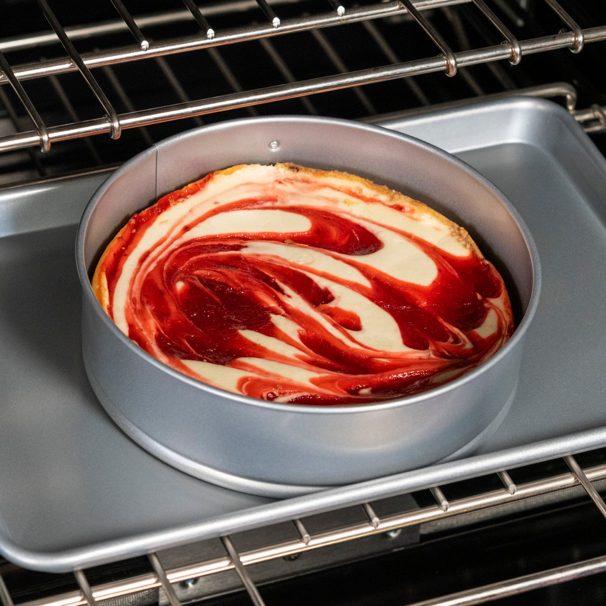 A round cheesecake with red and white swirled topping is baking in a Viking Culinary Products Aluminized Steel Nonstick 9-inch Springform Pan, set on a baking sheet inside an oven.
