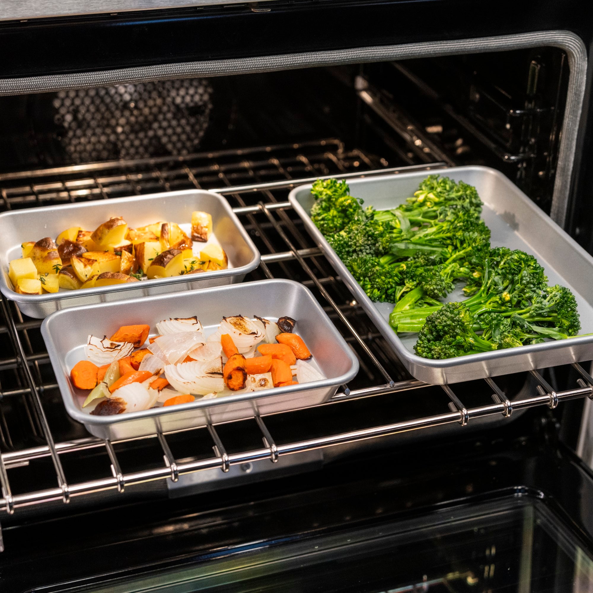 Three Viking Culinary Products Aluminized Steel Nonstick Nesting Baking Sheets, filled with chopped broccoli, carrots and onions, and yellow potatoes, roast together on an oven rack inside an open oven.