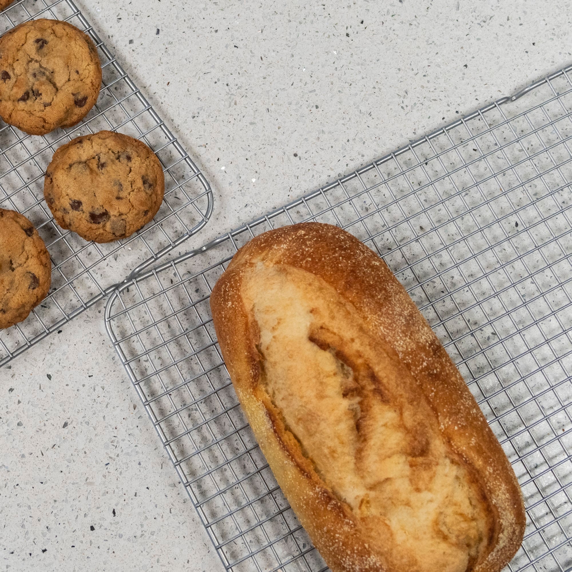 A loaf of bread and several chocolate chip cookies are cooling on the Viking Culinary Products 2-pc Cooling Rack Set (fits 17-inch baking sheet) atop a light-colored countertop.