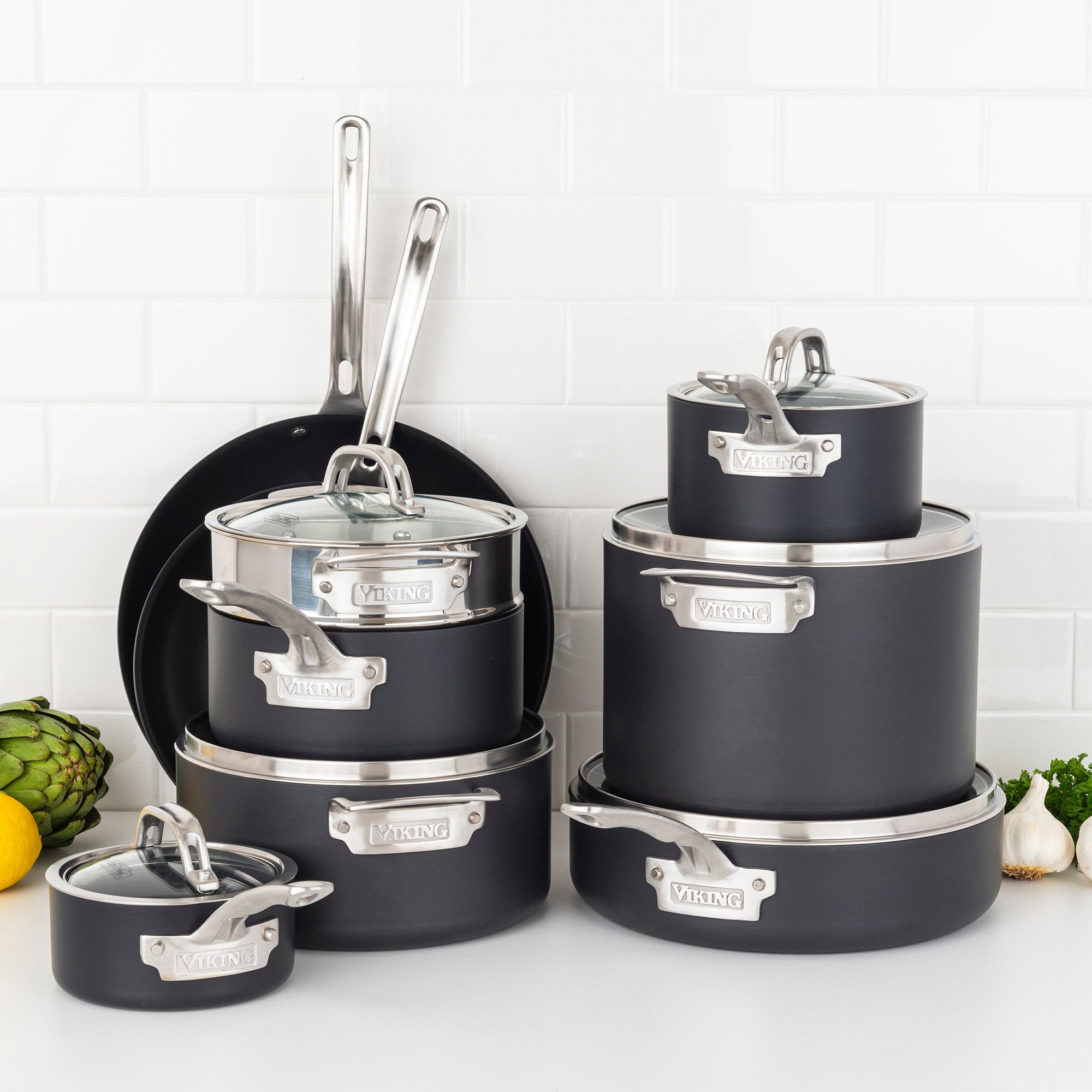The Viking Hard Anodized Nonstick 15-Piece Cookware Set with Glass Lids from Viking Culinary Products is arranged on a kitchen counter with a white tile backsplash, alongside artichoke, lemon, and garlic. The set is induction capable.