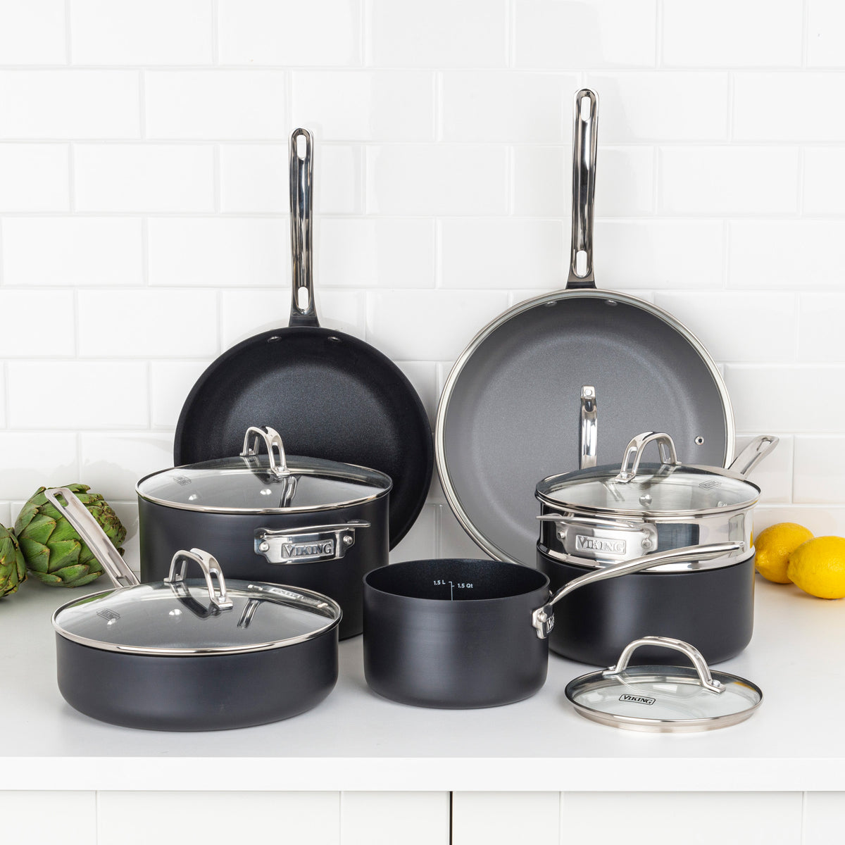 A Viking Culinary Products Hard Anodized Ceramic Nonstick 12pc Cookware Set, PFAS free and black with stainless steel handles and glass lids, is arranged on a white kitchen counter with lemons and an artichoke in the background.