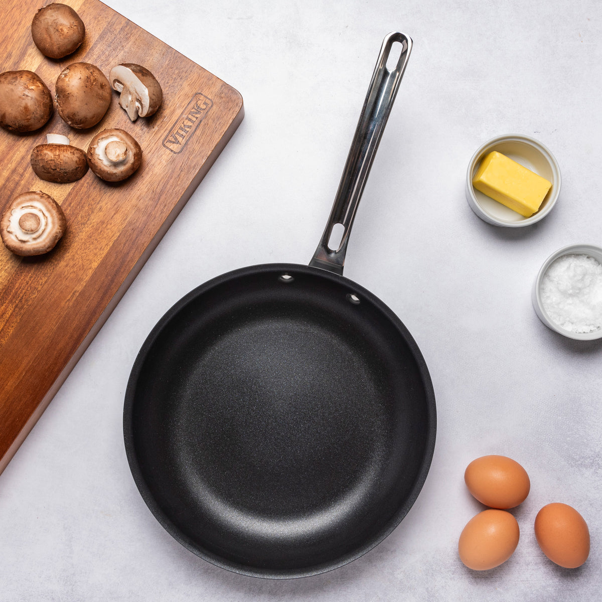 The Viking Hard Anodized Ceramic Nonstick 12pc Cookware Set by Viking Culinary Products sits on a light surface with a cutting board of sliced mushrooms, butter, salt, and three brown eggs nearby.