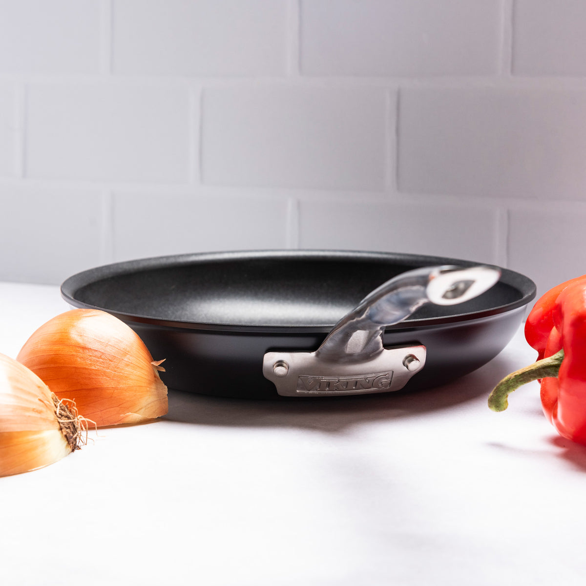 A Viking Hard Anodized Ceramic Nonstick 12pc Cookware pan by Viking Culinary Products rests on a white surface with two yellow onions to the left, a red bell pepper to the right, and PFAS free assurance shown against tiled walls.