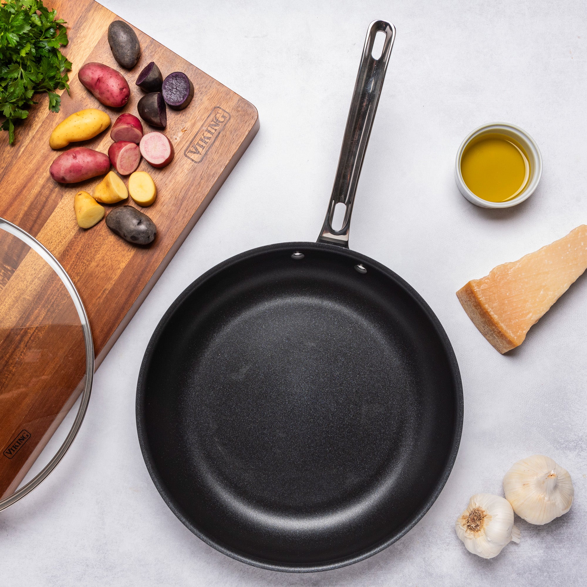 A black frying pan from the Viking Culinary Products Hard Anodized Ceramic Nonstick 12pc Cookware Set sits on a white surface, surrounded by a cutting board with chopped potatoes, parsley, cheese, garlic, and a bowl of olive oil.