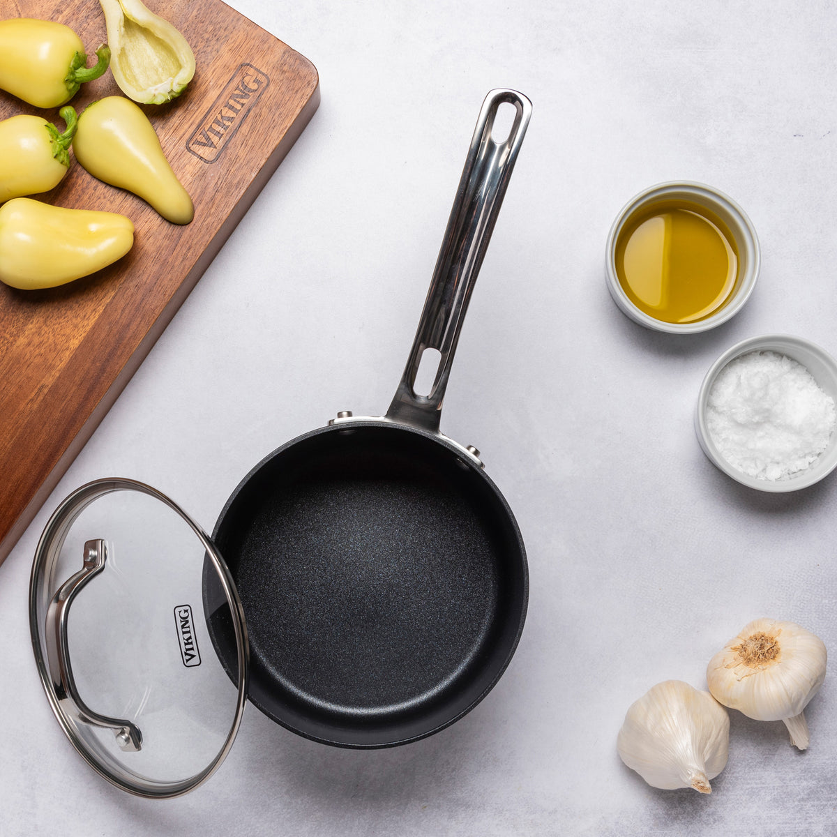 A small empty saucepan with lid from the Viking Hard Anodized Ceramic Nonstick 12pc Cookware Set by Viking Culinary Products sits on a white countertop, surrounded by yellow peppers, olive oil, salt, and garlic.