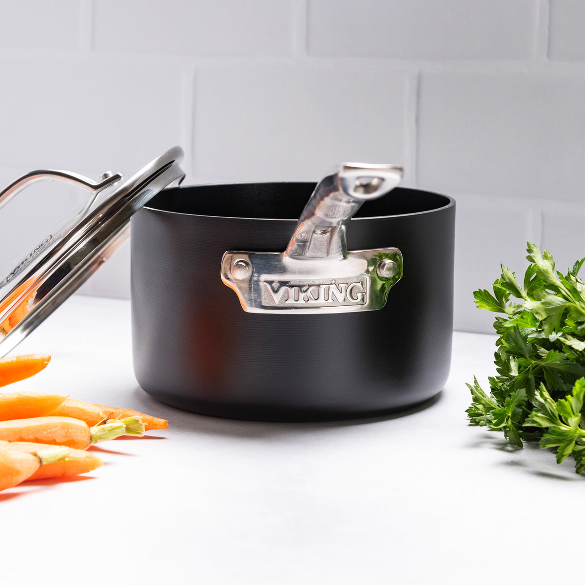 A Viking Hard Anodized Ceramic Nonstick saucepan from Viking Culinary Products, featuring a metal handle and lid, rests on a white countertop beside fresh carrots and parsley with a backdrop of white tiled walls.