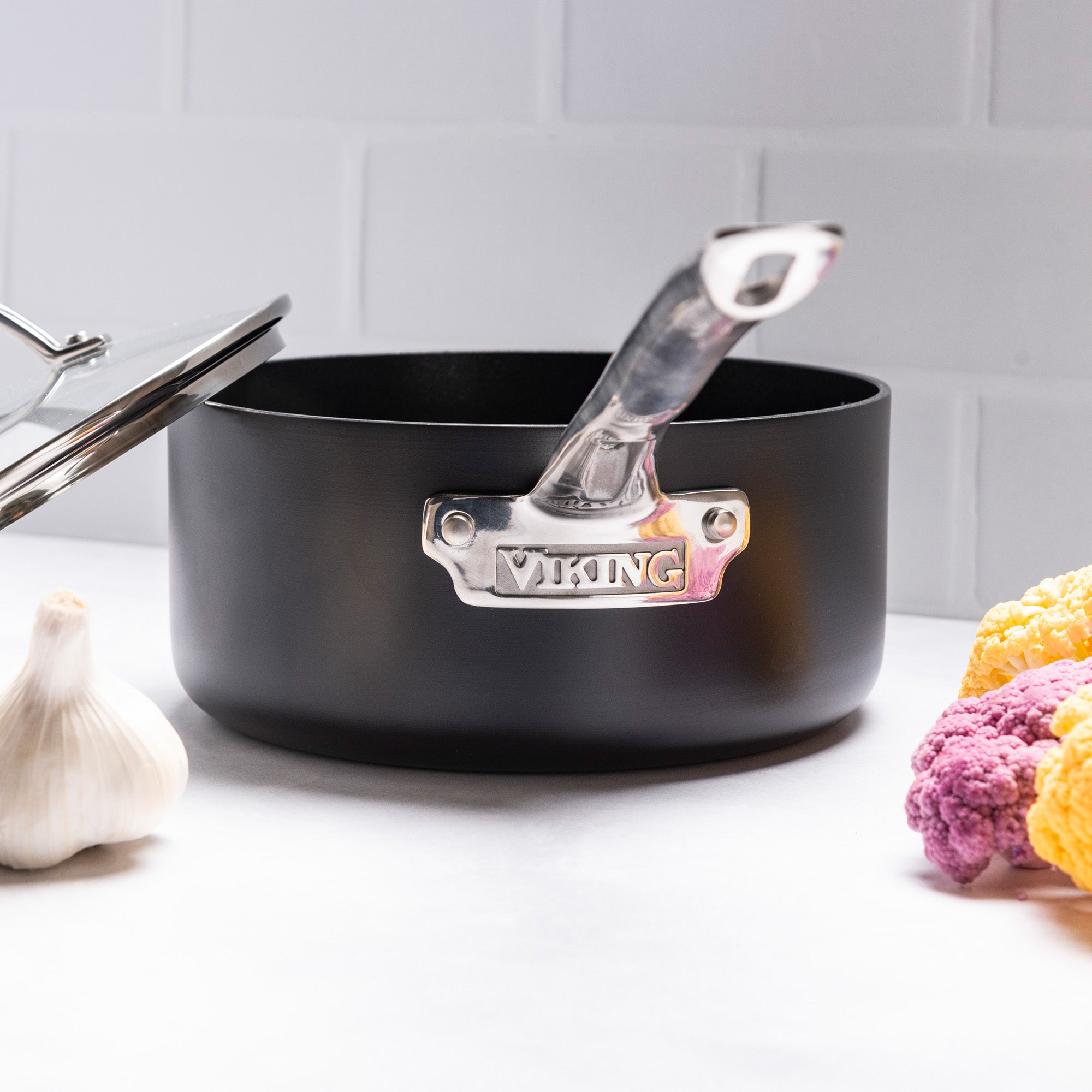 A black Viking Culinary Products hard anodized ceramic nonstick saucepan with metal handle and lid, part of the Viking Hard Anodized Ceramic Nonstick 12pc Cookware Set, sits on a white surface beside garlic and colorful cauliflower against white tiles. PFAS free.