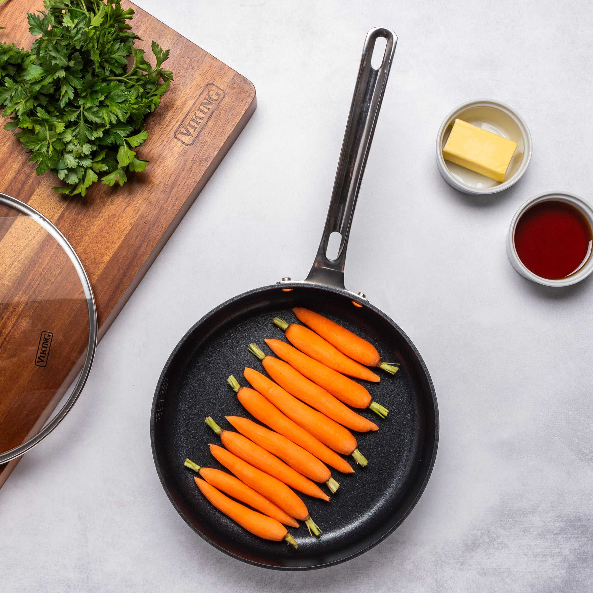 A Viking Hard Anodized Ceramic Nonstick pan from the Viking Culinary Products 12pc set holds baby carrots on a white surface, accompanied by parsley, butter, a glass lid, and red liquid—ideal for any PFAS-free kitchen.