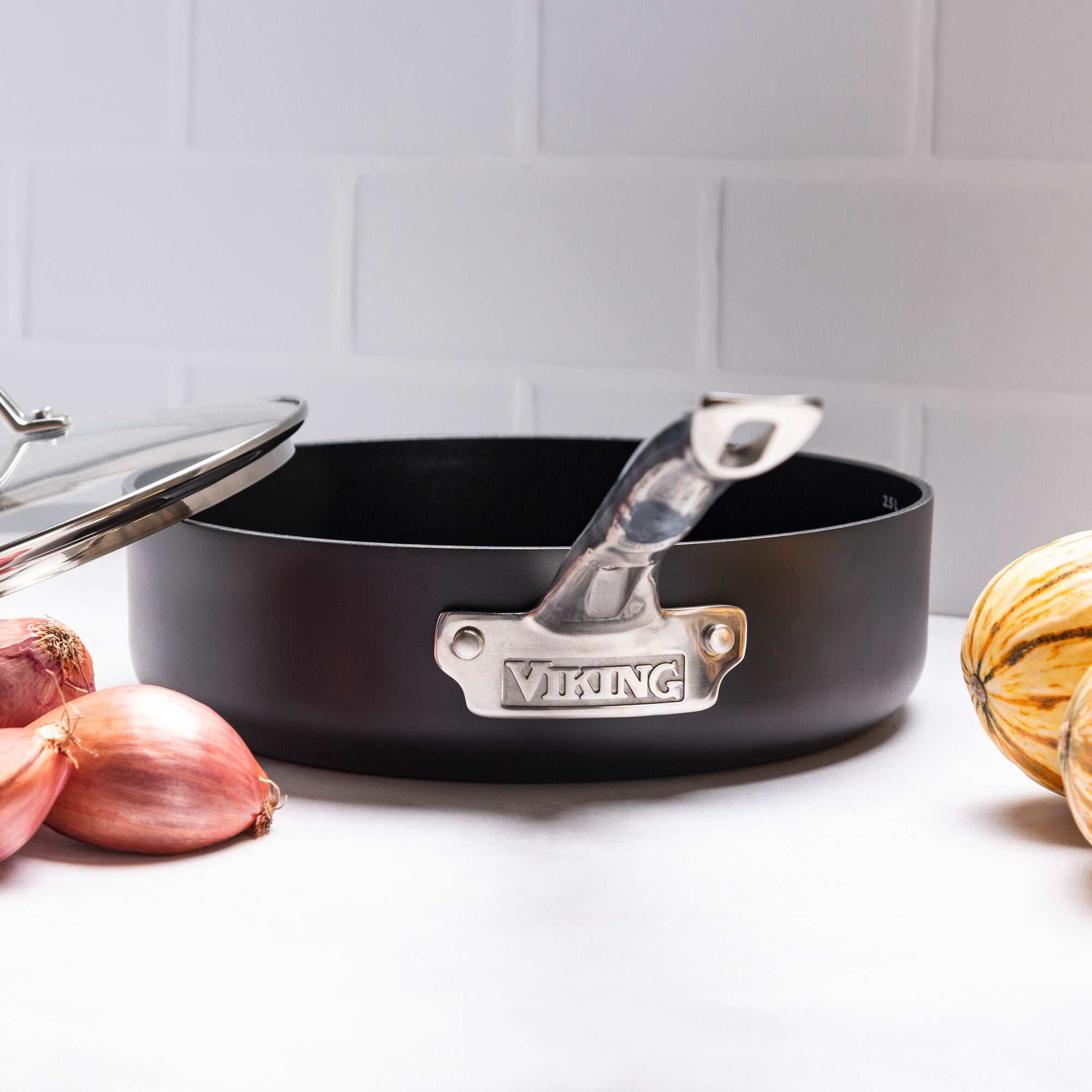 A black skillet from the Viking Culinary Products Hard Anodized Ceramic Nonstick 12pc Cookware Set, featuring a metal handle and lid, sits on a white surface with shallots and striped squash, against a white tiled background.