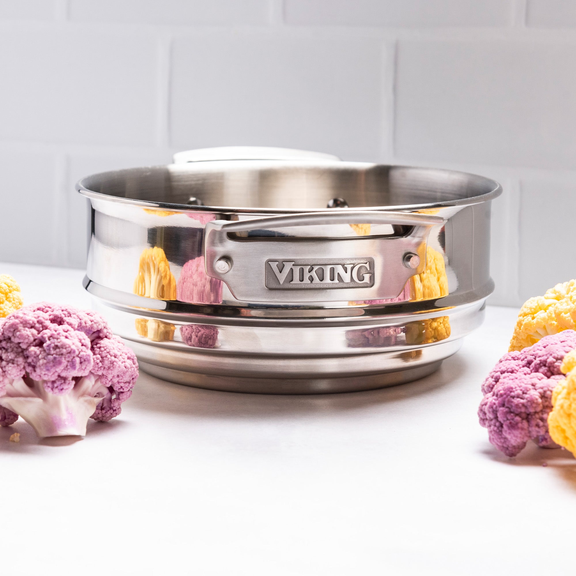 A Viking Culinary Products Hard Anodized Ceramic Nonstick 12pc Cookware set steamer basket rests on a white surface with purple and yellow cauliflower florets, set against a white tiled wall.