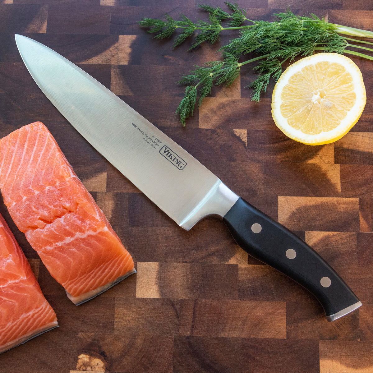 A chef’s knife on a wooden cutting board next to two pieces of raw salmon, a halved lemon, and sprigs of fresh dill.