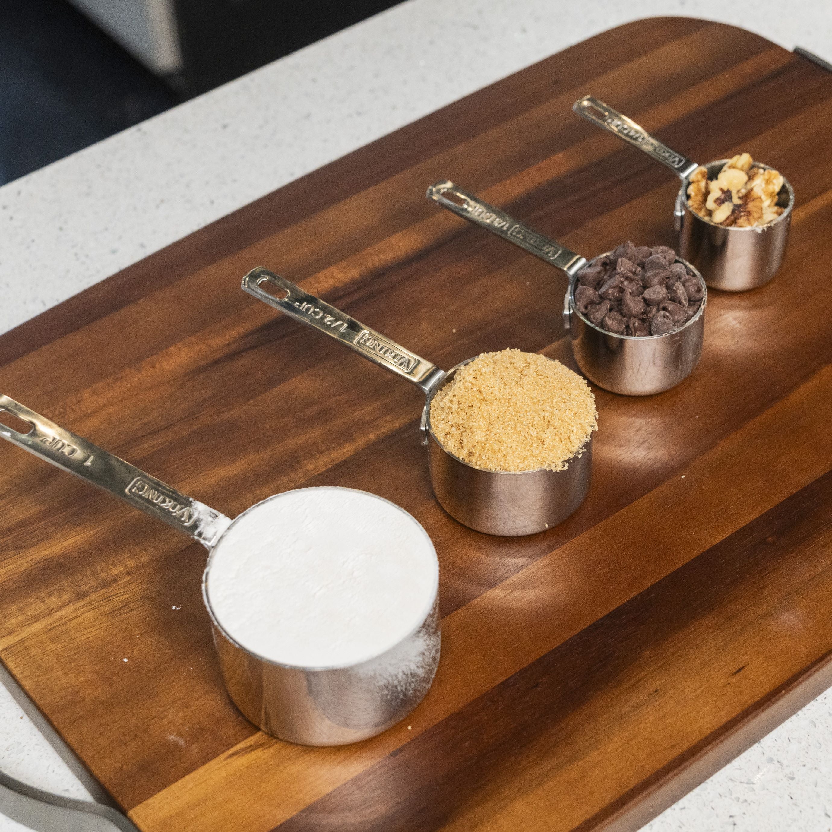 The Viking Stainless Steel Measuring Cup Set by Viking Culinary Products is arranged on a wooden board from front to back, each cup filled with flour, brown sugar, chocolate chips, and chopped walnuts.