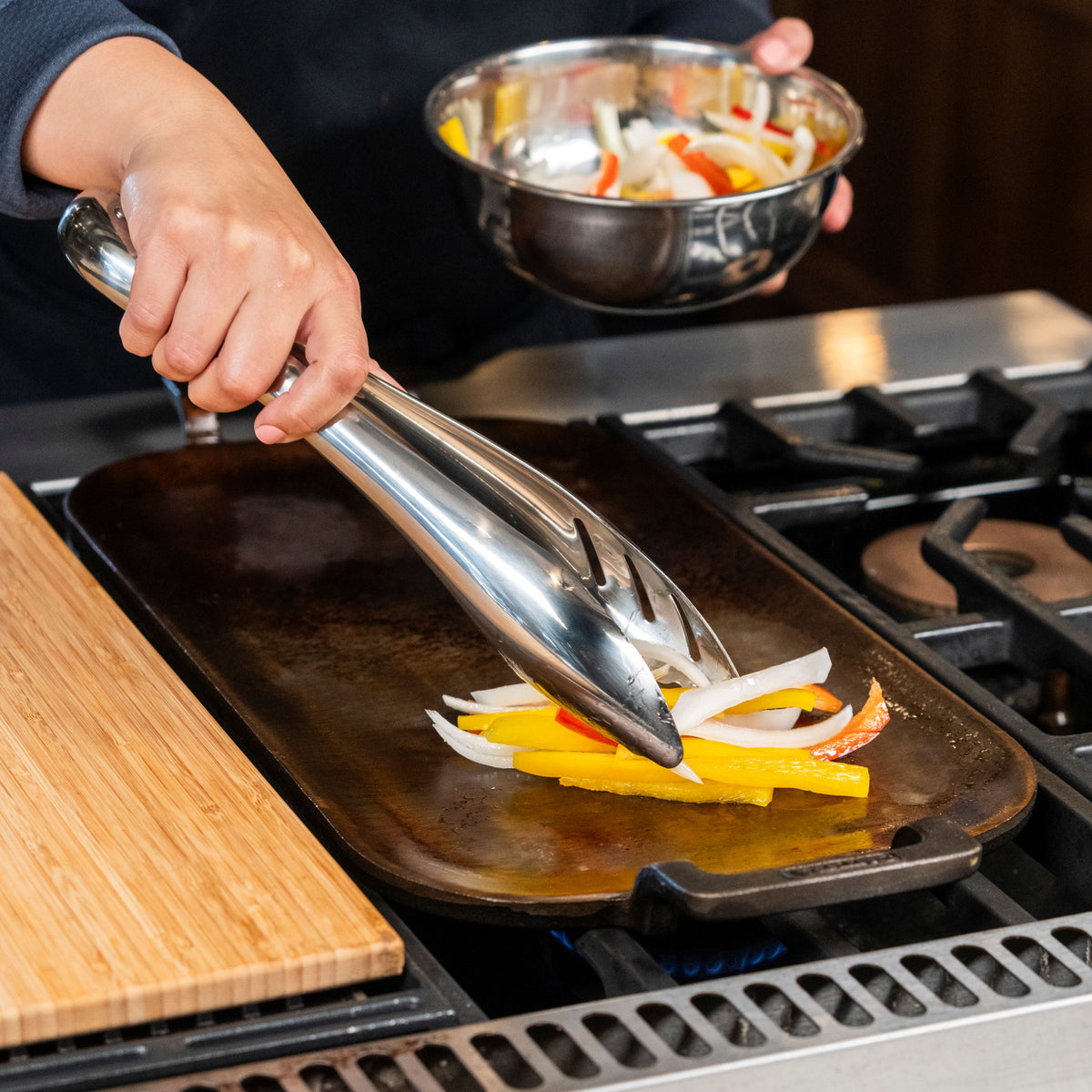 Using Viking 12-inch Stainless Steel Gravity Tongs by Viking Culinary Products, a person transfers sliced yellow and red bell peppers and onions onto a hot griddle, while keeping extra vegetables in a metal bowl nearby for easy cleanup.
