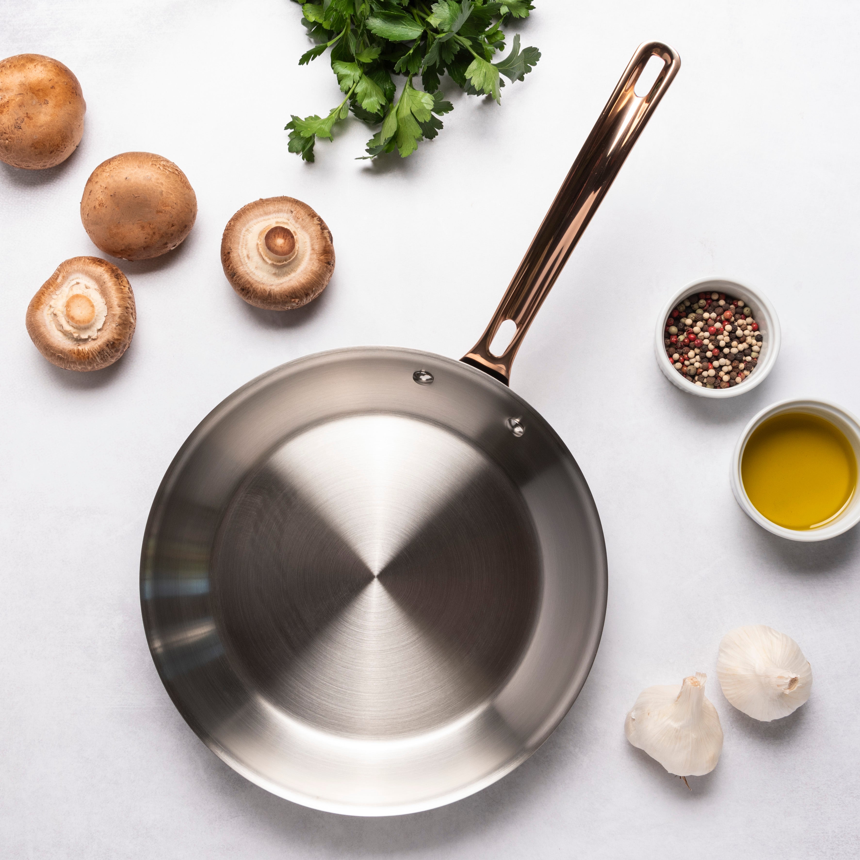 The Viking 10pc 3-Ply Black & Copper Stainless Steel Cookware Set by Viking Culinary Products features a PFAS-free frying pan with copper handle, surrounded by mushrooms, parsley, peppercorns, olive oil, and garlic on a white surface—ideal for safe, stylish cooking.