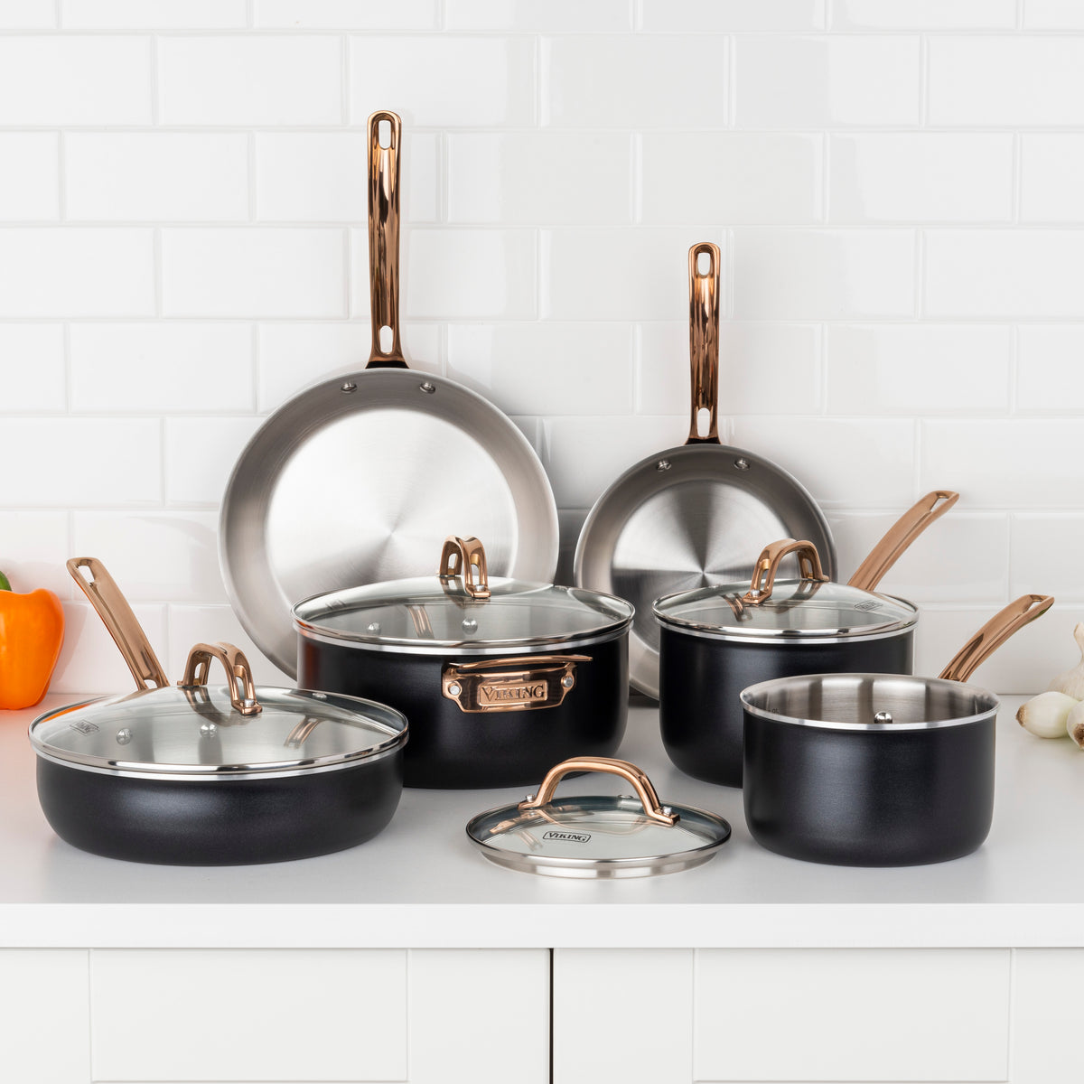 The Viking 10pc 3-Ply Black & Copper Stainless Steel Cookware Set by Viking Culinary Products is displayed on a white counter with glass-lidded pots, copper-colored handles, and an orange bell pepper on the left.