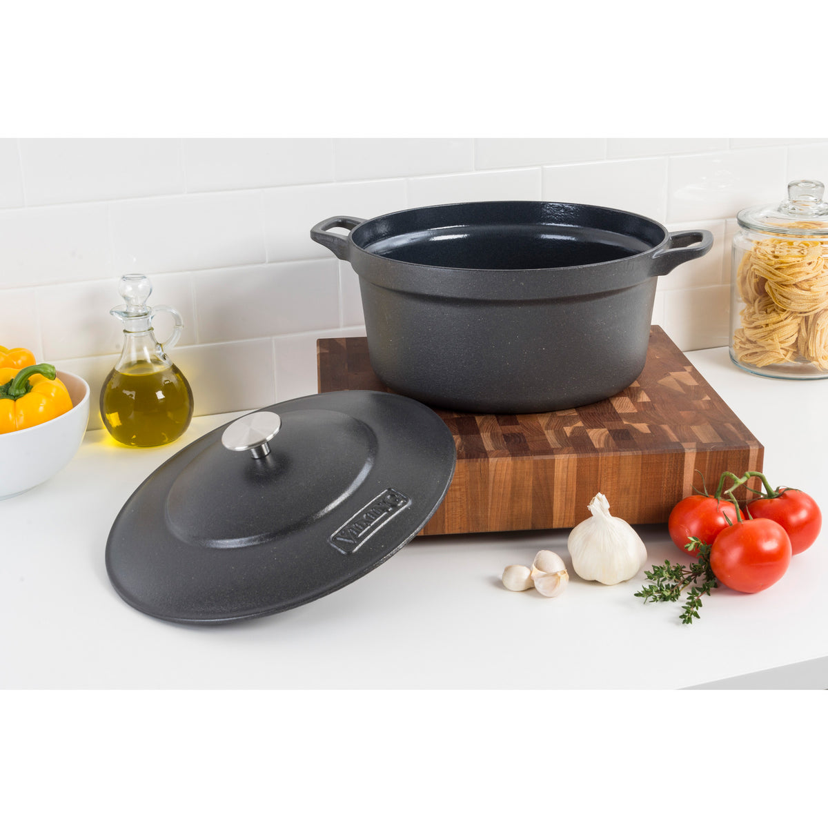A black cast iron pot with a lid sits on a wooden cutting board. Nearby are tomatoes, garlic, thyme, olive oil, a yellow bell pepper, and a jar of pasta on a white kitchen counter.