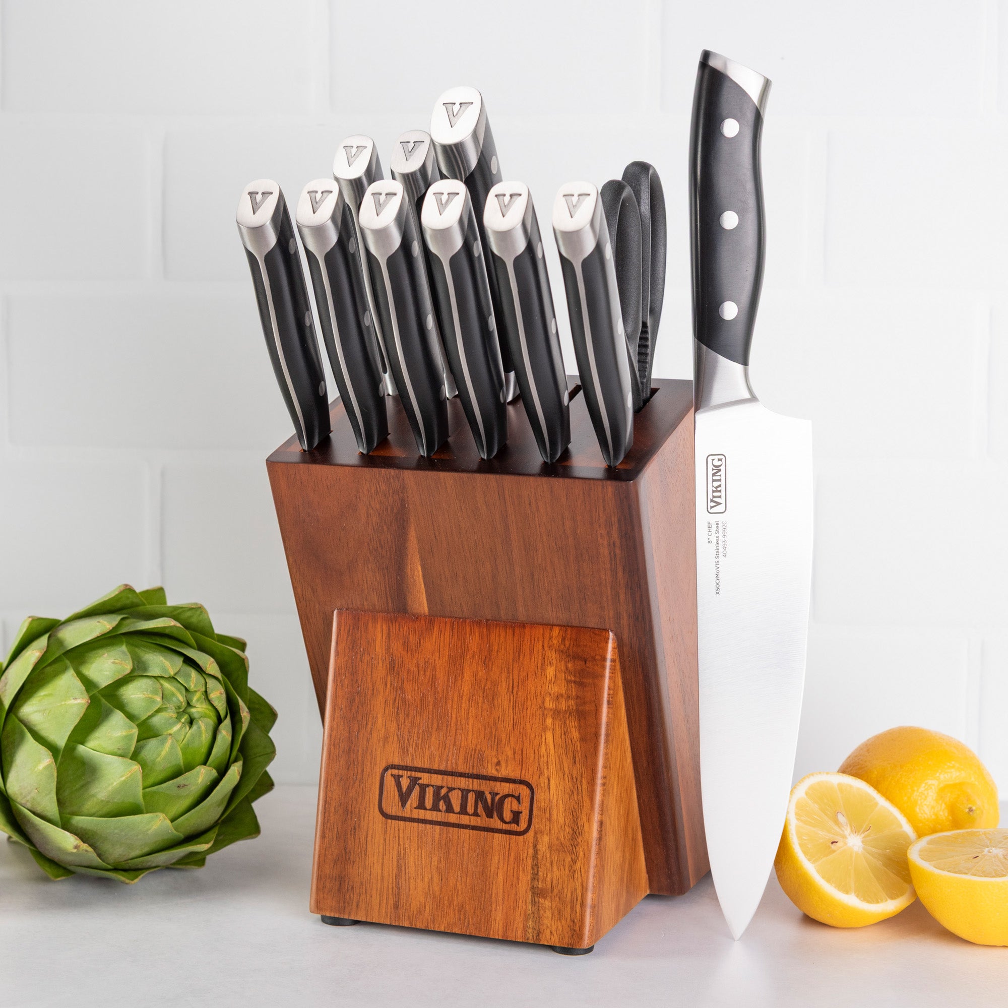 The Viking 12-Piece Stainless Steel Forged Cutlery Block Set by Viking Culinary Products features high carbon knives with black handles and silver tips, including steak and chef’s knives, displayed on a counter near an artichoke and sliced lemons.