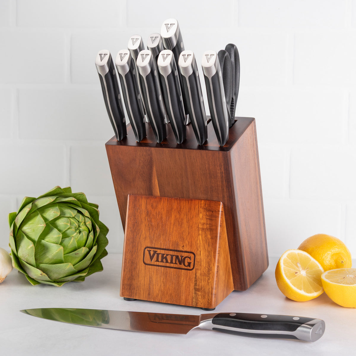 A Viking Culinary Products 12-Piece Stainless Steel Forged Cutlery Block Set holds knives and kitchen shears. A chef's knife rests on the counter by a halved lemon, whole lemon, and artichoke, with a white tiled wall behind.