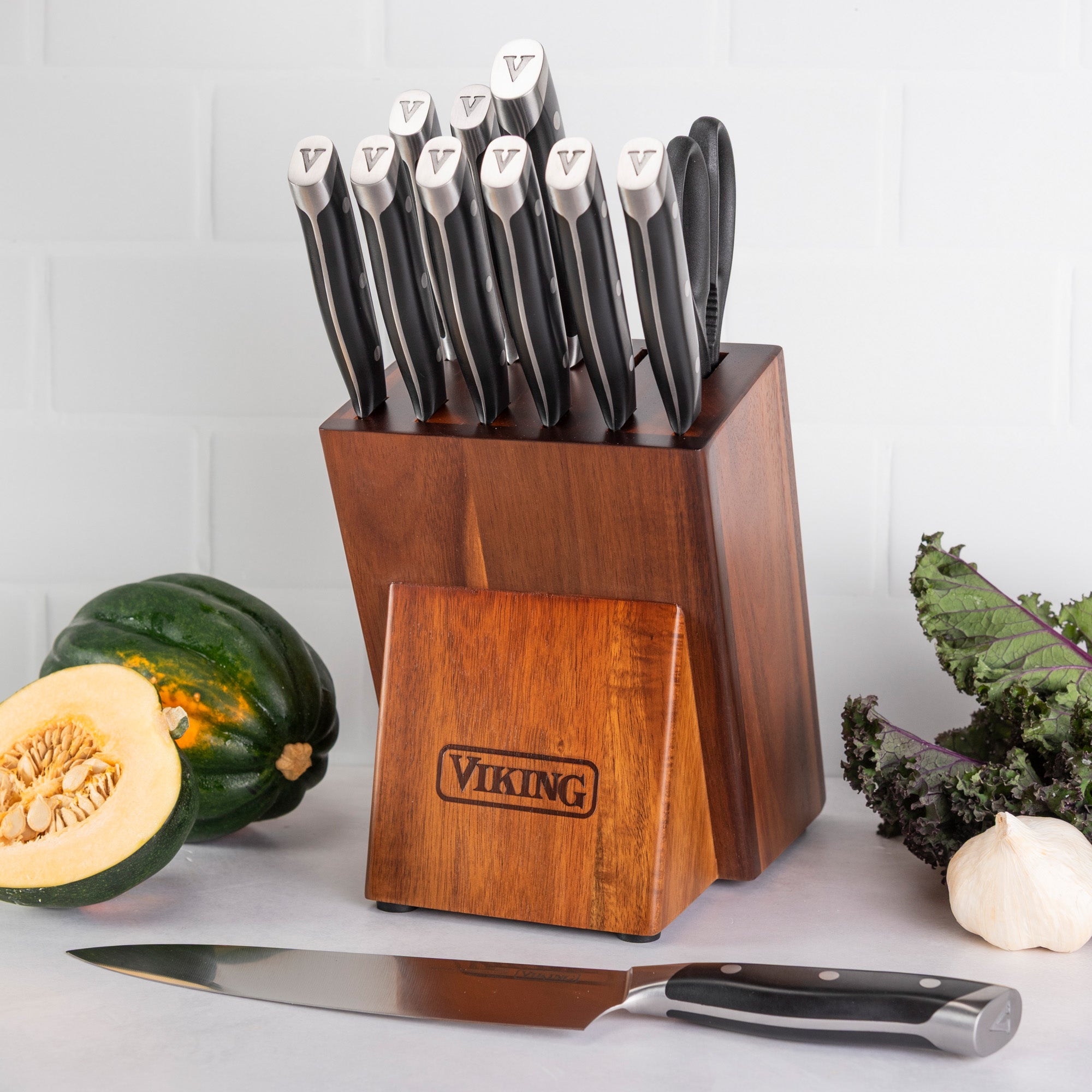 A Viking Culinary Products 12-piece stainless steel forged cutlery block set holds knives with silver and black handles. A chef’s knife rests by a halved squash, leafy greens, and garlic on the counter.