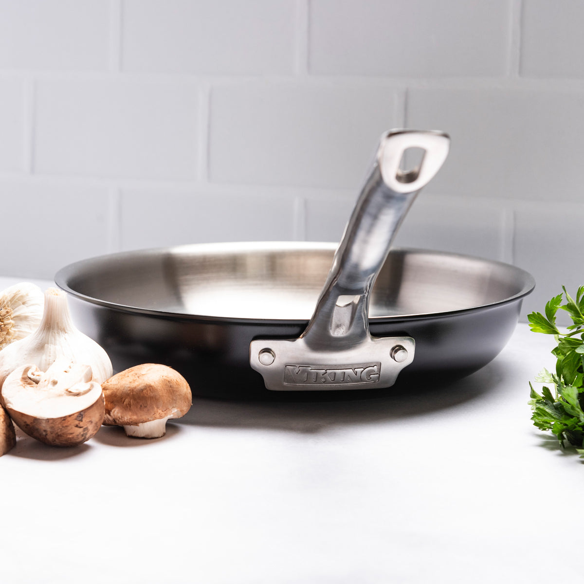 The Viking Culinary Products 12pc 3-Ply Cast Black Exterior Stainless Steel Cookware Set with Metal Lids is shown on a white surface with mushrooms, garlic, and parsley nearby. Induction compatible and PFAS free, with riveted handles and polished interiors.
