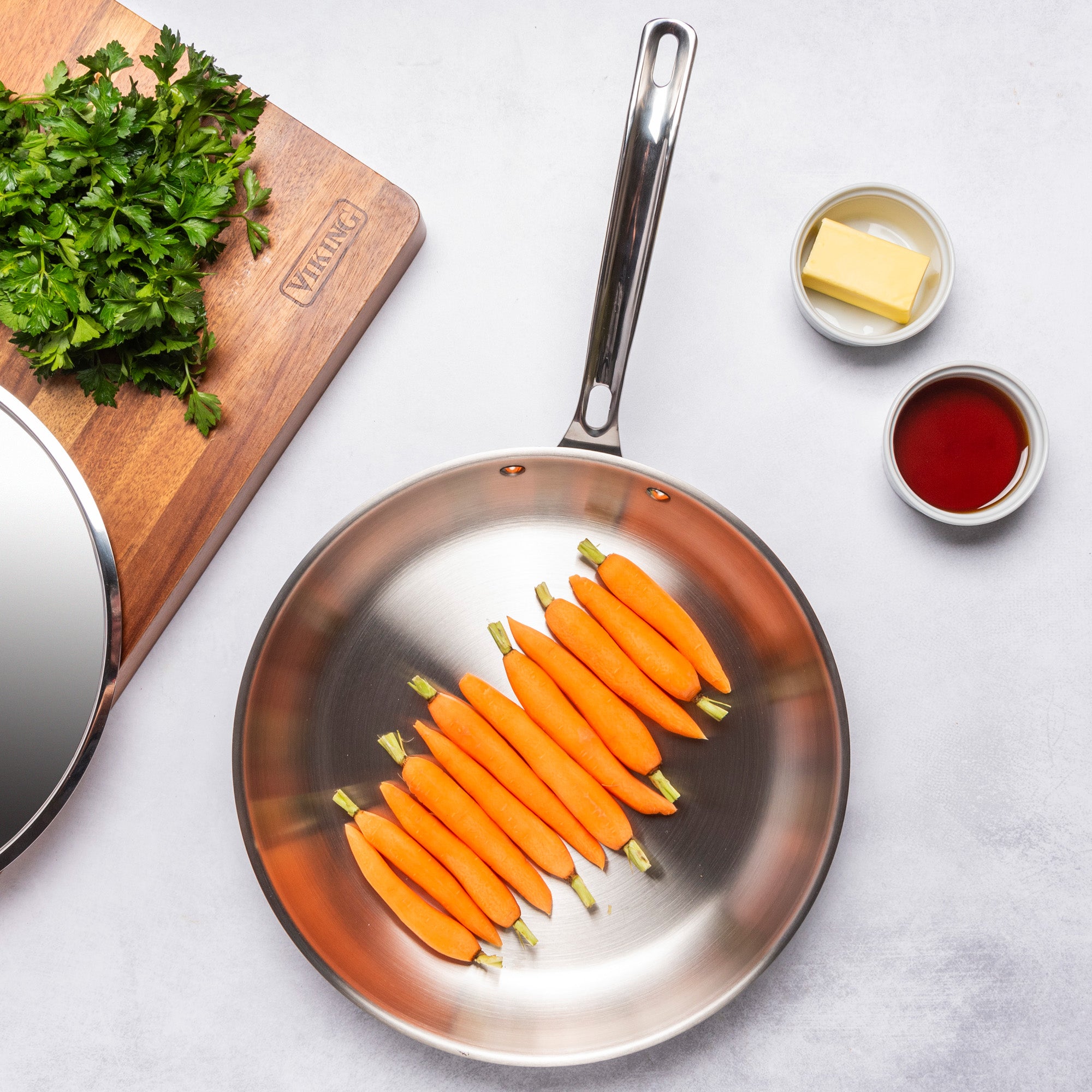 A PFAS-free, induction-compatible Viking 12pc 3-Ply Cast Black Exterior Stainless Steel pan by Viking Culinary Products holds peeled baby carrots beside a wooden board with chopped parsley, butter, and a bowl of dark liquid on a light countertop.