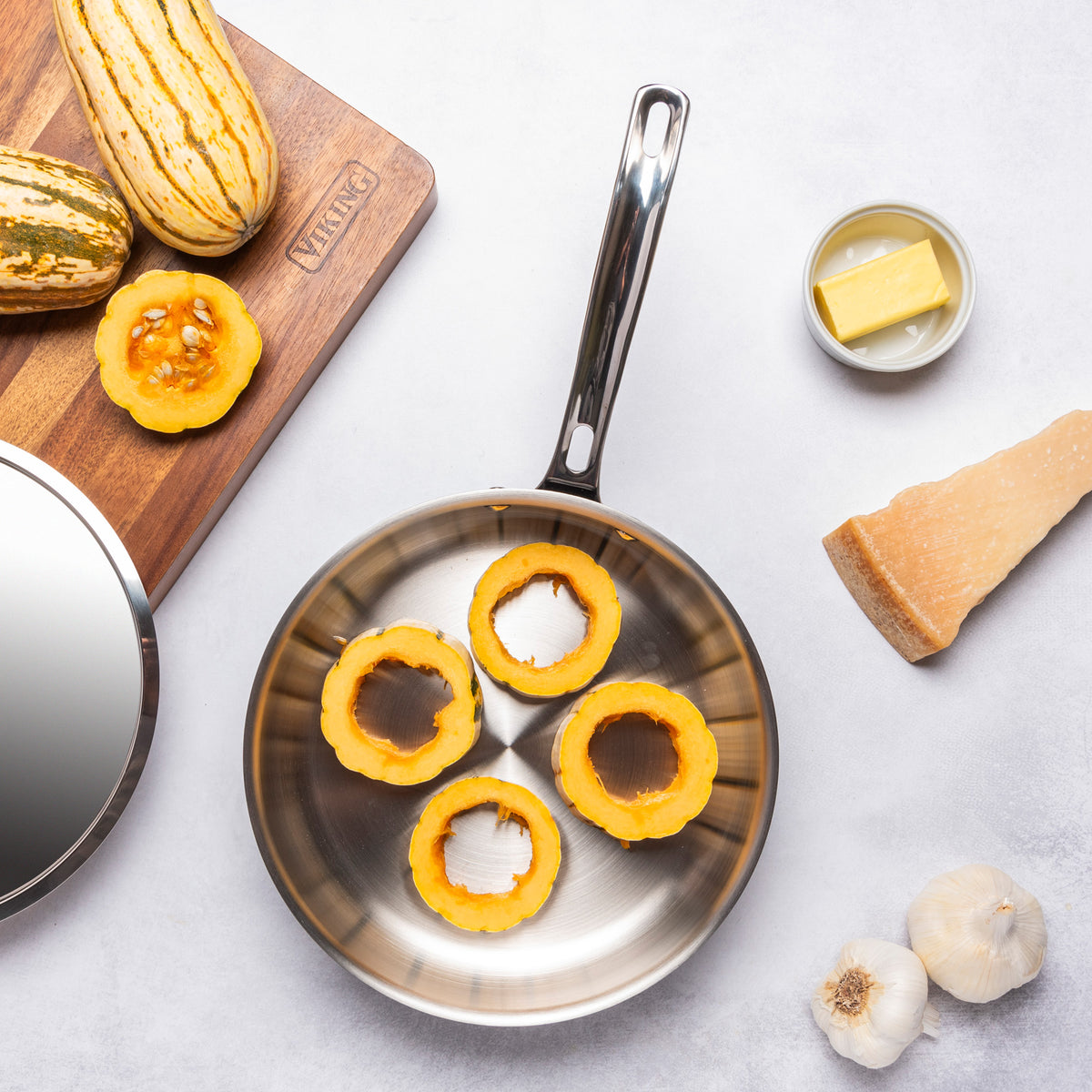 A Viking Culinary Products 12pc 3-Ply Cast Black Exterior Stainless Steel pan from their cookware set sits on a white surface with four raw squash rings, surrounded by whole squash, a cutting board, Parmesan cheese, garlic bulbs, and butter.