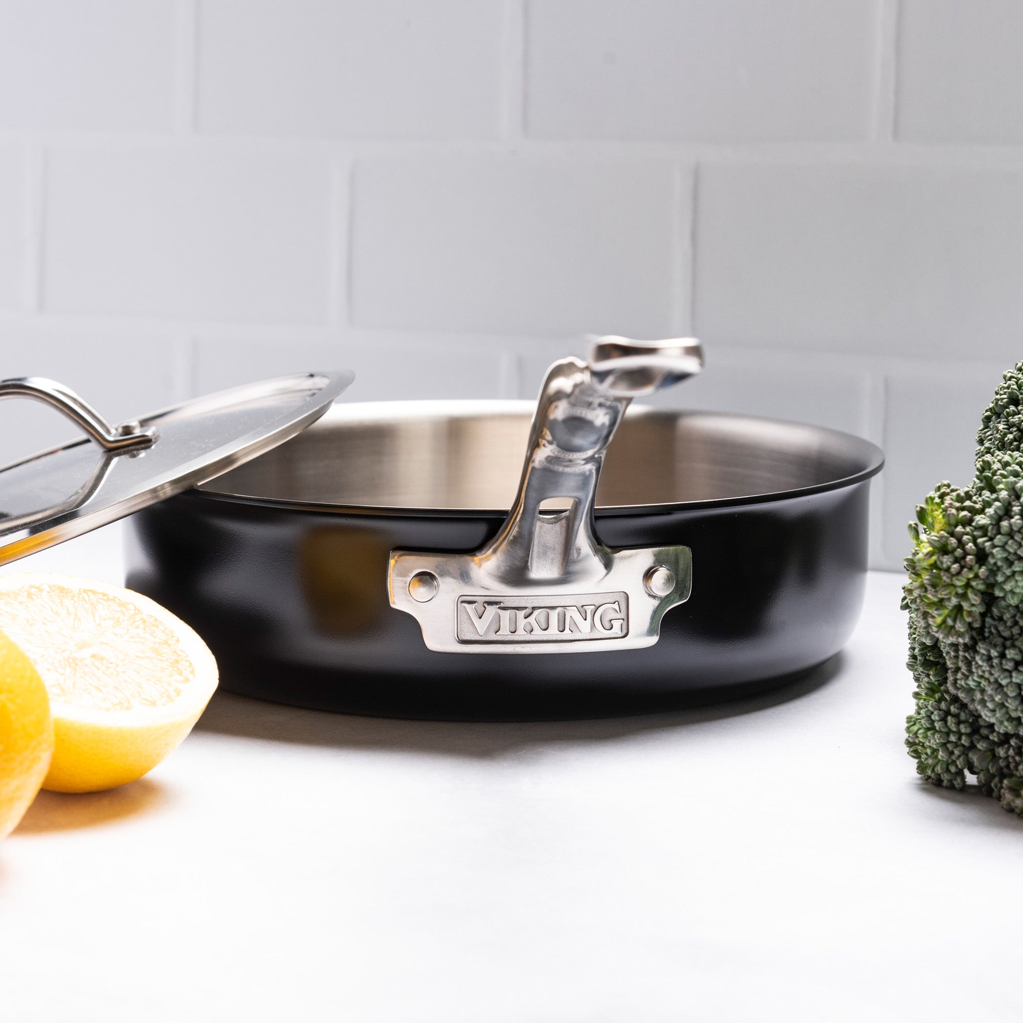 A PFAS-free Viking Culinary Products 12pc 3-Ply Cast Black Exterior Stainless Steel Cookware Set with metal lids sits on a white countertop near halved lemons and broccoli—ideal for induction cooking, against a white tiled background.