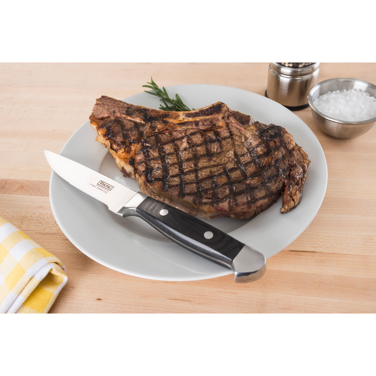 A grilled steak with grill marks sits on a white plate with a steak knife. A sprig of rosemary, a salt bowl, salt and pepper shakers, and a yellow plaid napkin are nearby on a wooden table.