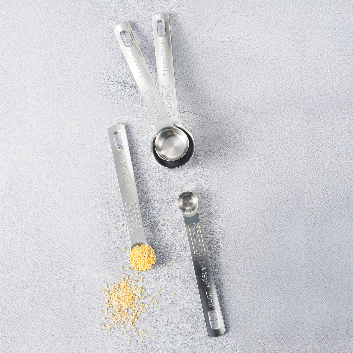 The Viking Culinary Products 4pc Stainless Steel Measuring Spoon Set is arranged on a light grey surface, with one spoon holding grains and others stacked empty. These spoons are dishwasher safe for easy cleaning.
