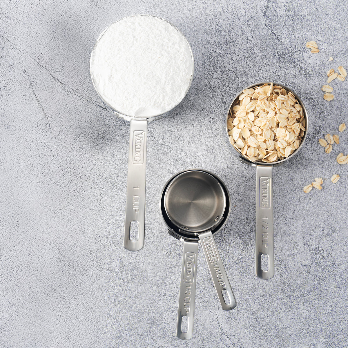 The Viking Stainless Steel Measuring Cup Set by Viking Culinary Products, labeled from 1 cup to 1/4 cup, is filled with flour and oats on a gray surface with scattered oats. These dishwasher safe cups make baking easy and tidy.