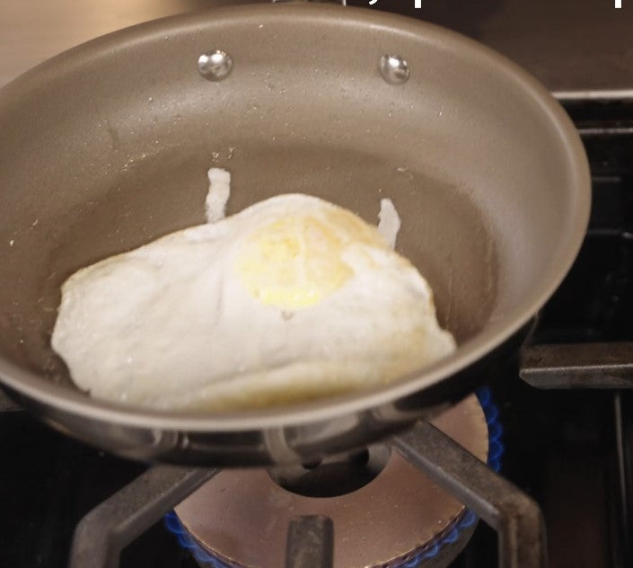A fried egg cooks in a Viking PureGlide Pro 3-Ply 8 Inch Fry Pan by Viking Culinary Products on a gas stove, with the burner flame visible beneath the pan.