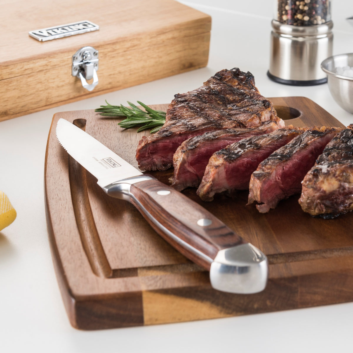 A sliced grilled steak rests on a wooden cutting board with a steak knife beside it. A sprig of rosemary, a wooden box, a pepper grinder, and a metal bowl are in the background.