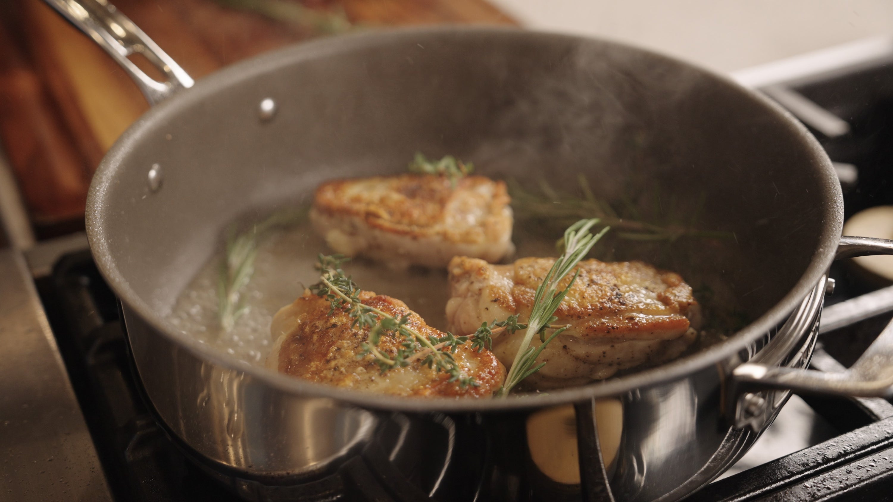 Three pieces of golden-brown chicken cook with rosemary and thyme, steam rising, in a Viking PureGlide Pro 3-Ply 4.5-Quart Saute Pan with Glass Lid from Viking Culinary Products on the stovetop.